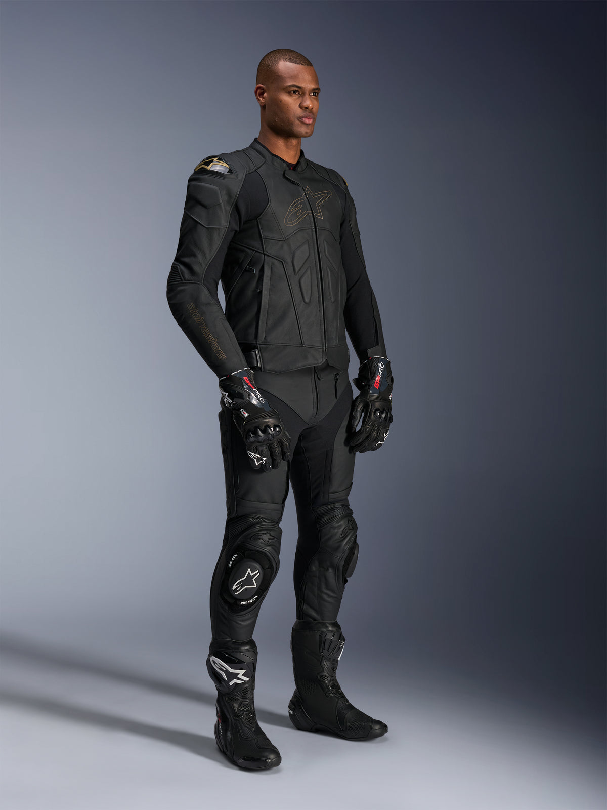 Mannelijk model in Alpinestars Missile V3 leren broek en bijpassende zwarte leren jas met gouden accenten, zwarte GP Pro racehandschoenen en zwarte racelaarzen, complete motorrace-uitrusting voor weggebruik, studio-opname.