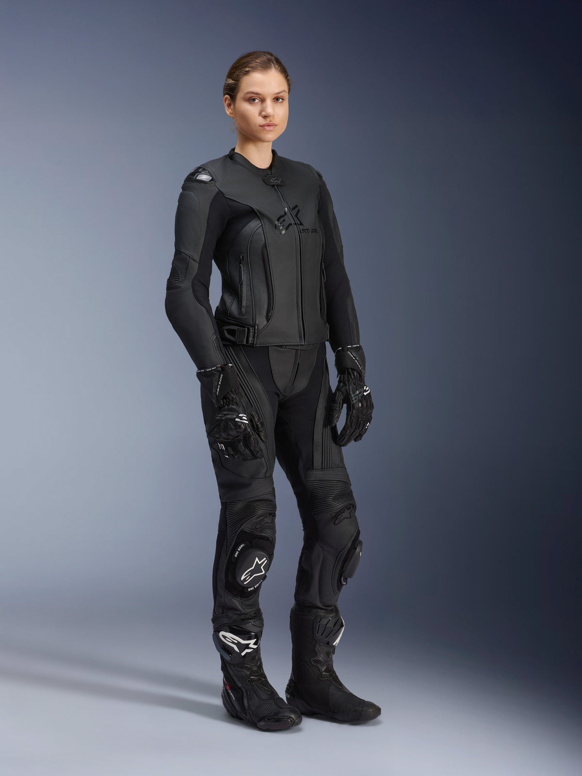 Vrouwelijke motorrijder draagt de Alpinestars Stella GP Plus V4 leren damesjas in Black Black, gestyled als een volledig leren racepak met bijpassende motorbroek, handschoenen en laarzen, voorzien van een gestroomlijnd performance-ontwerp met geïntegreerde bescherming.