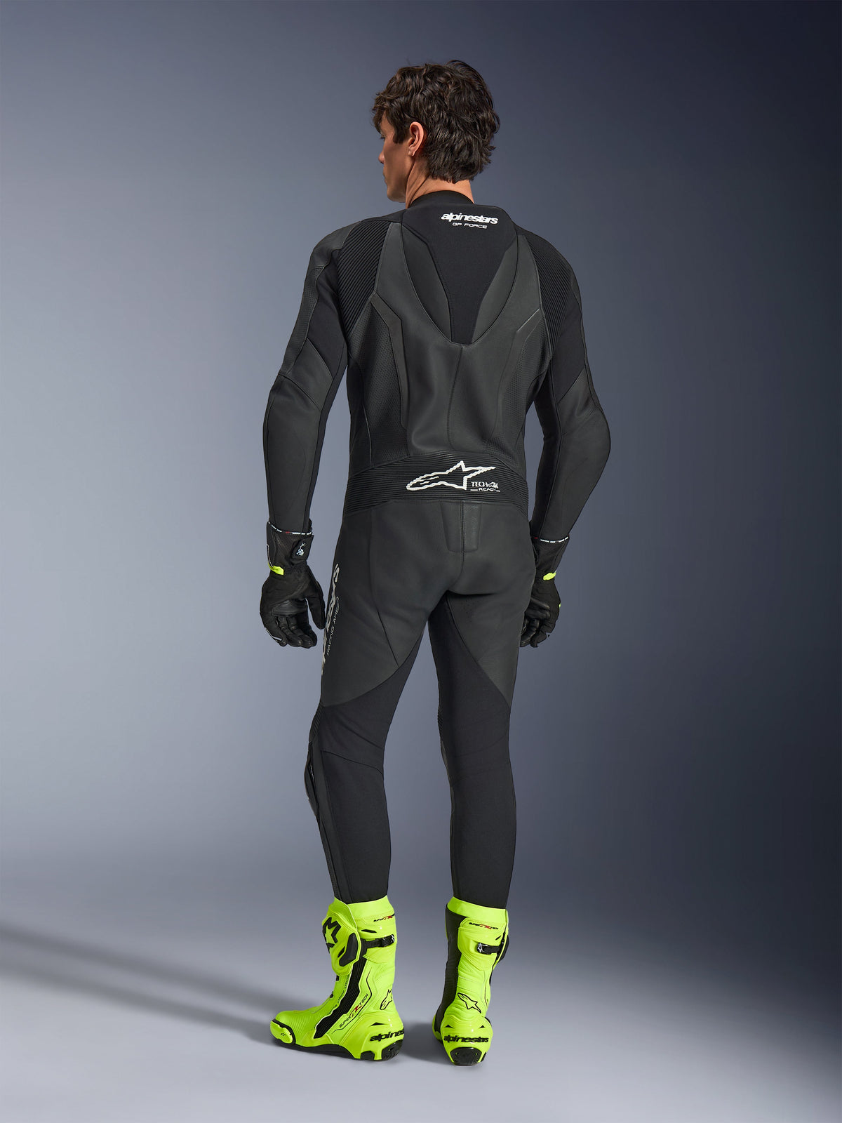 Alpinestars GP Force V2 Leren Pak - 1-delig, zwart, achteraanzicht gedragen door een model, voorzien van een aerodynamische speedhump en Tech-Air Ready branding, gecombineerd met zwarte handschoenen en fel fluorescerende gele racelaarzen, professionele circuituitrusting