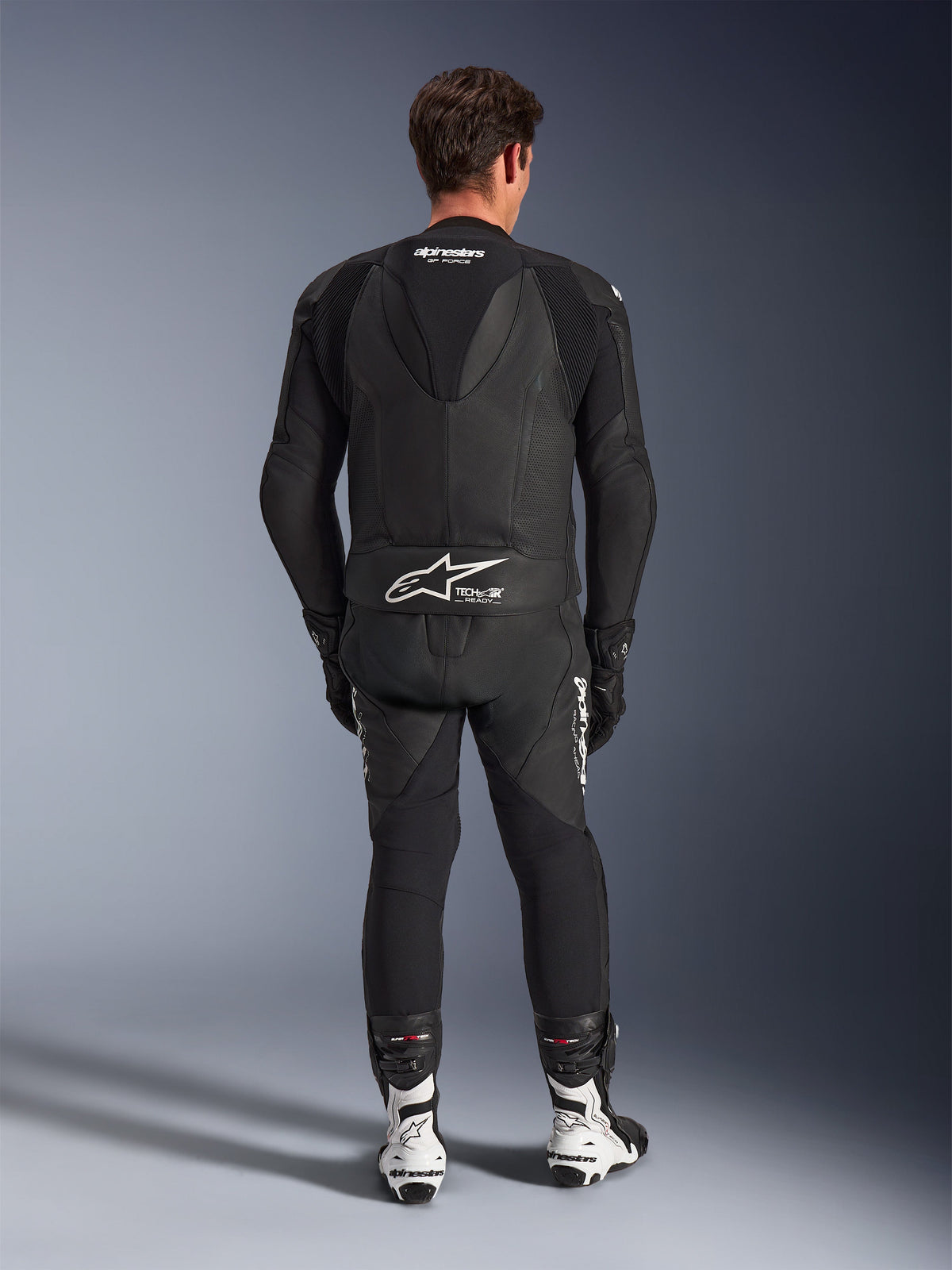 Alpinestars GP Force V2 tweedelig leren pak, zwart, achteraanzicht met aerodynamische speedhump en Tech-Air Ready-branding op de taille, geperforeerde leren panelen voor ventilatie, gecombineerd met witte en zwarte racelaarzen, professionele motorrace-uitrusting