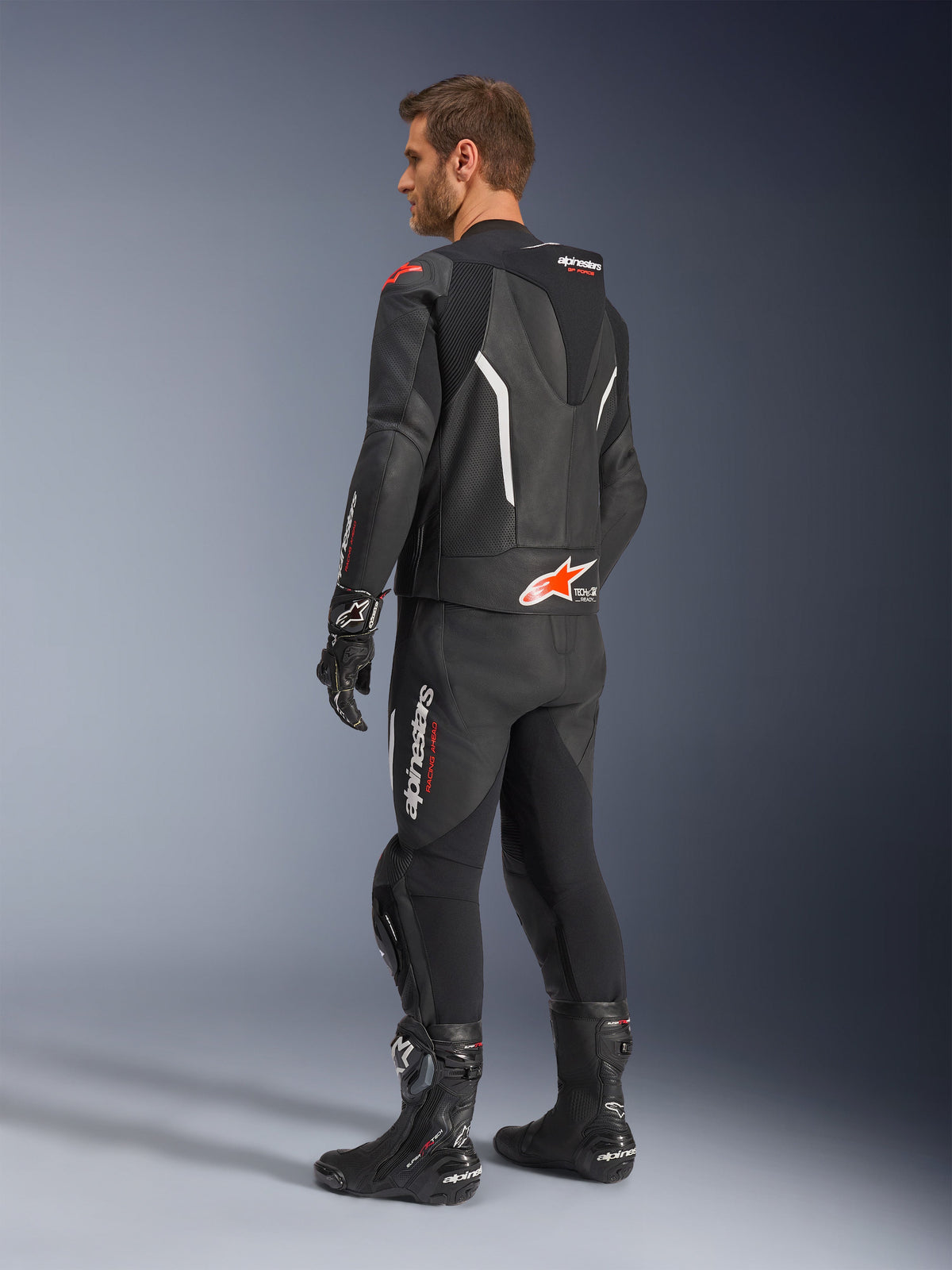 Alpinestars GP Force V2 tweedelig leren pak, zwart met witte en Red Fluo accenten, driekwart achteraanzicht op model, met een aerodynamische speed hump, geperforeerde leren panelen en opvallende Alpinestars branding, gedragen met zwarte racelaarzen en handschoenen.
