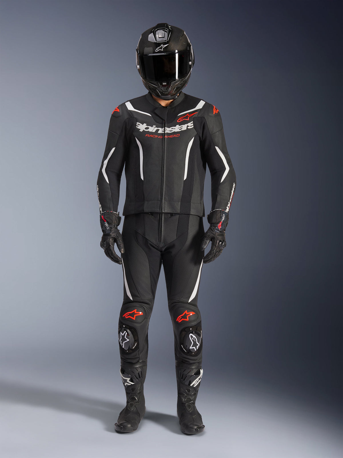 Alpinestars GP Force V2 tweedelig leren pak, motorracepak, zwart wit en rood fluo, voorzien van wit Alpinestars-logo op de borst en rode accenten op de schouders en knieën, gedragen met zwarte integraalhelm, zwarte handschoenen en zwarte laarzen voor een complete performance-uitrusting