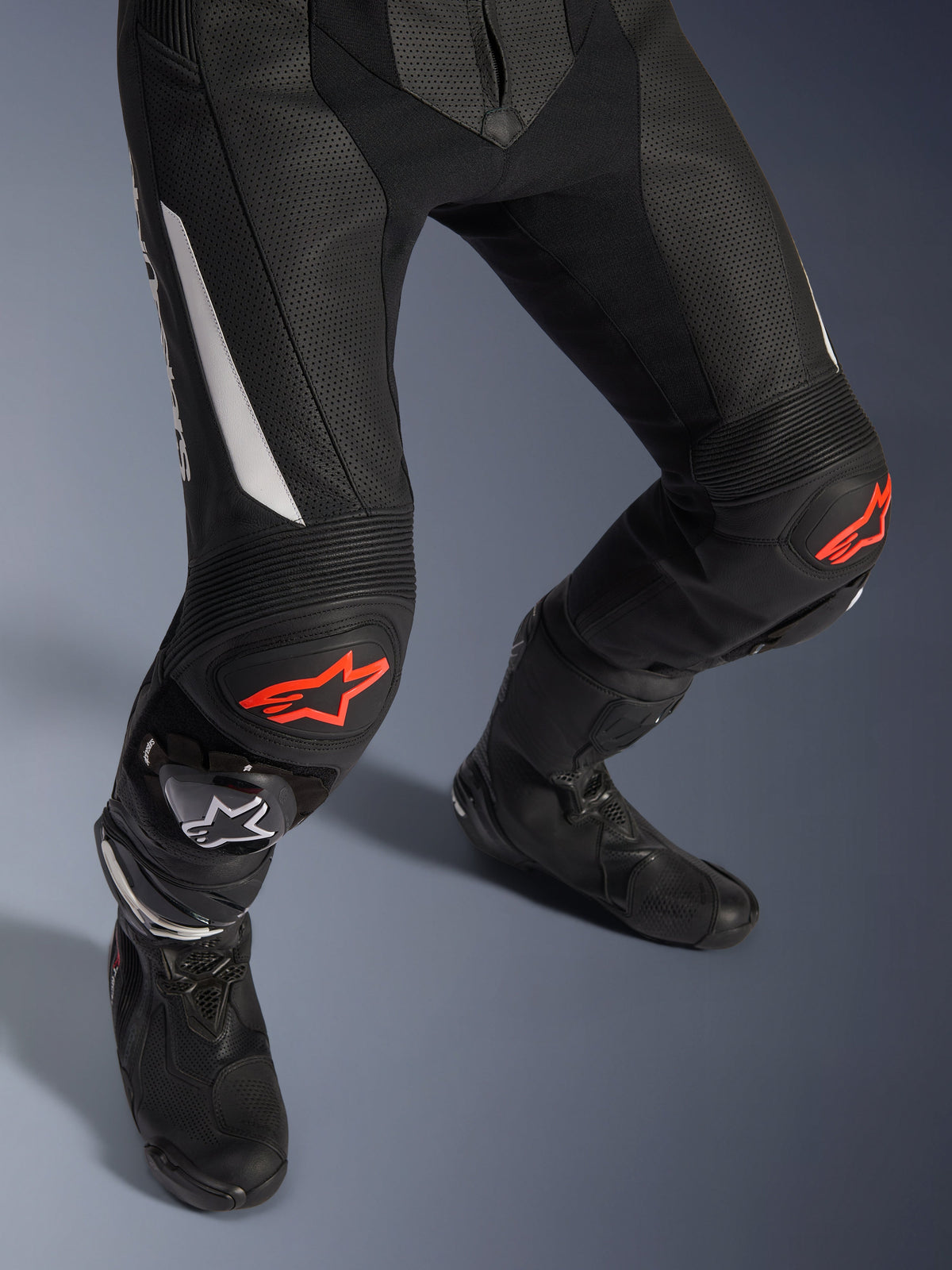 Alpinestars GP Force V2 2-delig leren pak, motorracebroek in zwart met rode en witte accenten, voorzien van geperforeerd leer, accordeon-stretchpanelen boven de knieën met rode sterlogo's, gedragen met zwarte motorlaarzen, hoogwaardige race-uitrusting