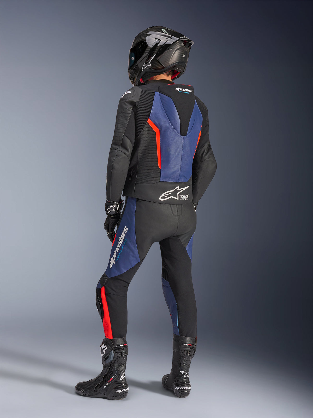 Alpinestars GP Force V2 tweedelig leren motorpak, zwart blauw rood fluo, achteraanzicht van rijder in professionele race-uitrusting, voorzien van een geïntegreerde speedhump en Tech-Air Ready branding, blauwe en rood fluo accenten op zwart leer, gedragen met een carbon fiber helm en zwarte laarzen, hoogwaardige motorkleding