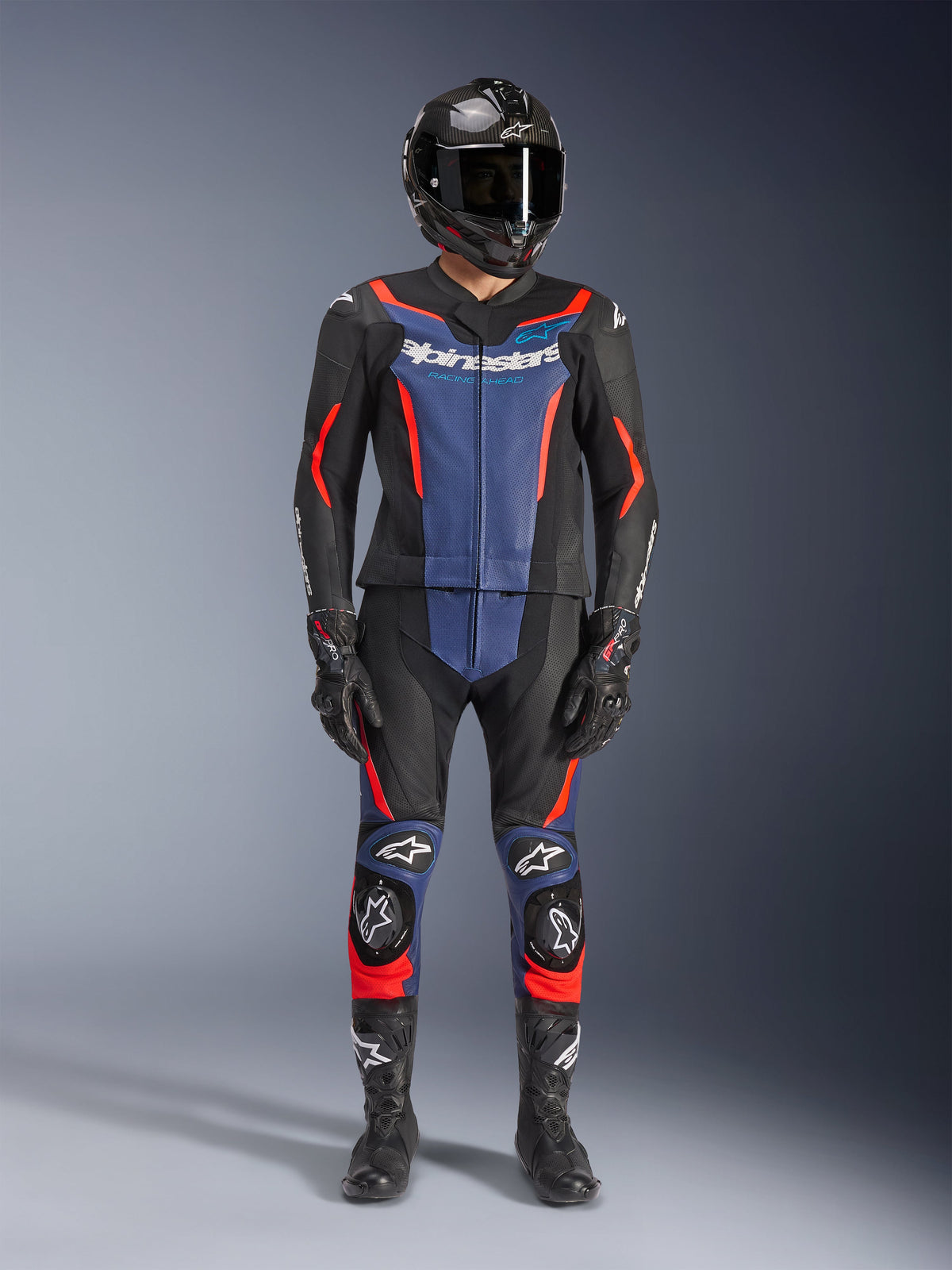Alpinestars GP Force V2 leren pak - 2-delig, zwart, blauw en rood fluo, gedragen met een carbon-fiber integraalhelm, zwarte racehandschoenen en laarzen, voorzien van blauwe borstpanelen en rood fluorescerende accenten, professionele motorrace-uitrusting
