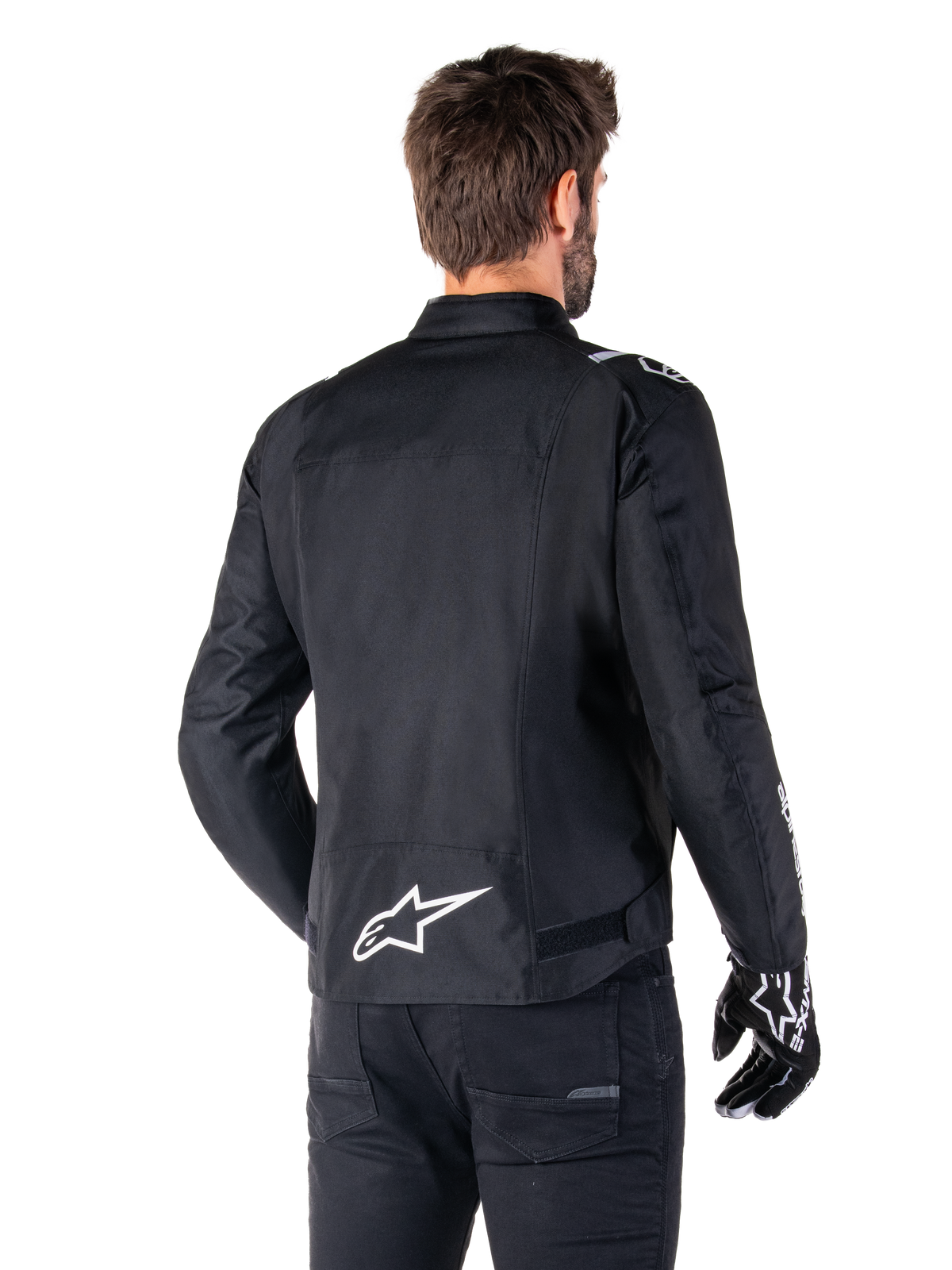 Alpinestars T-SPS V2 Waterproof Jacket, zwart en wit, achteraanzicht van een mannelijke rijder die de jas draagt, wit Alpinestars-logo op de onderrug, sportief gesneden textielconstructie, gedragen met zwarte motorbroek en handschoenen