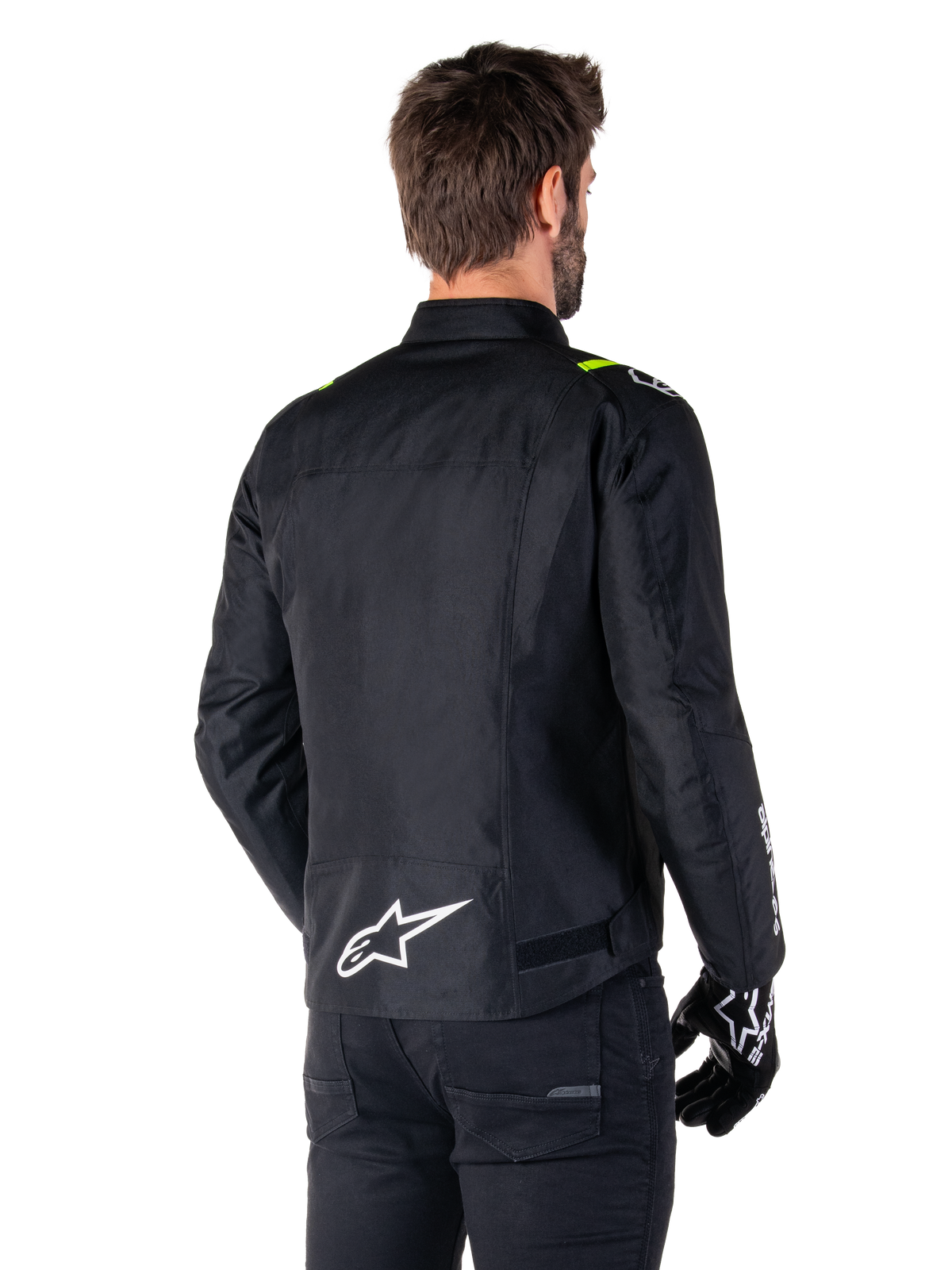 Alpinestars T-SPS V2 Waterproof Jacket, zwart wit fluogeel, achteraanzicht van een mannelijk model dat de textiele motorjas draagt, met een wit logo op de onderrug en fluogele accenten op de schouders, gecombineerd met een zwarte broek en handschoenen.