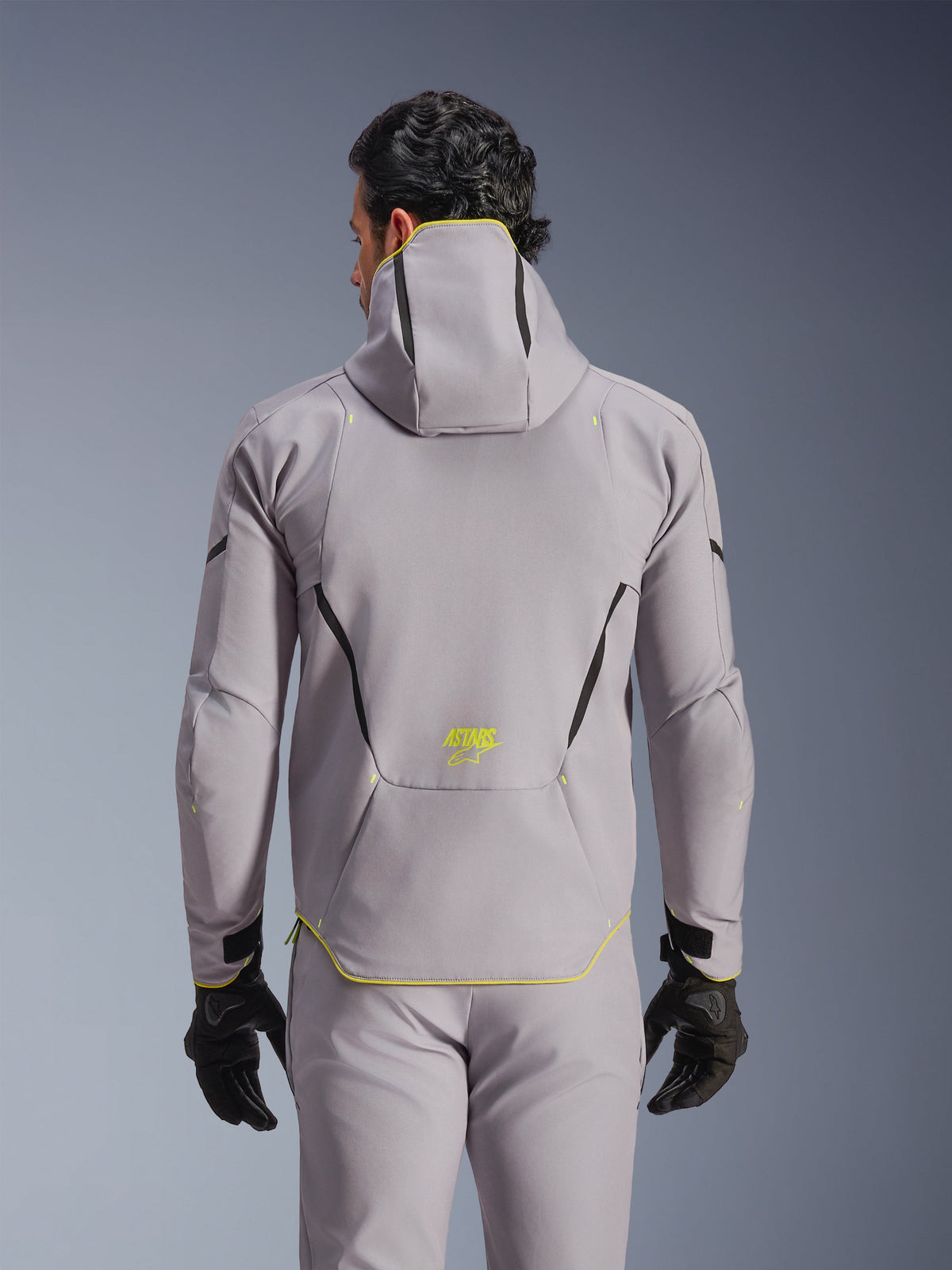 Alpinestars Aeroshell Waterproof Jacket, Mid Gray with Yellow Fluo accents, back view showing integrated hood and black contrast lines, yellow ASTARS logo on lower back, worn with matching gray pants and black gloves, sport softshell motorcycle jacket