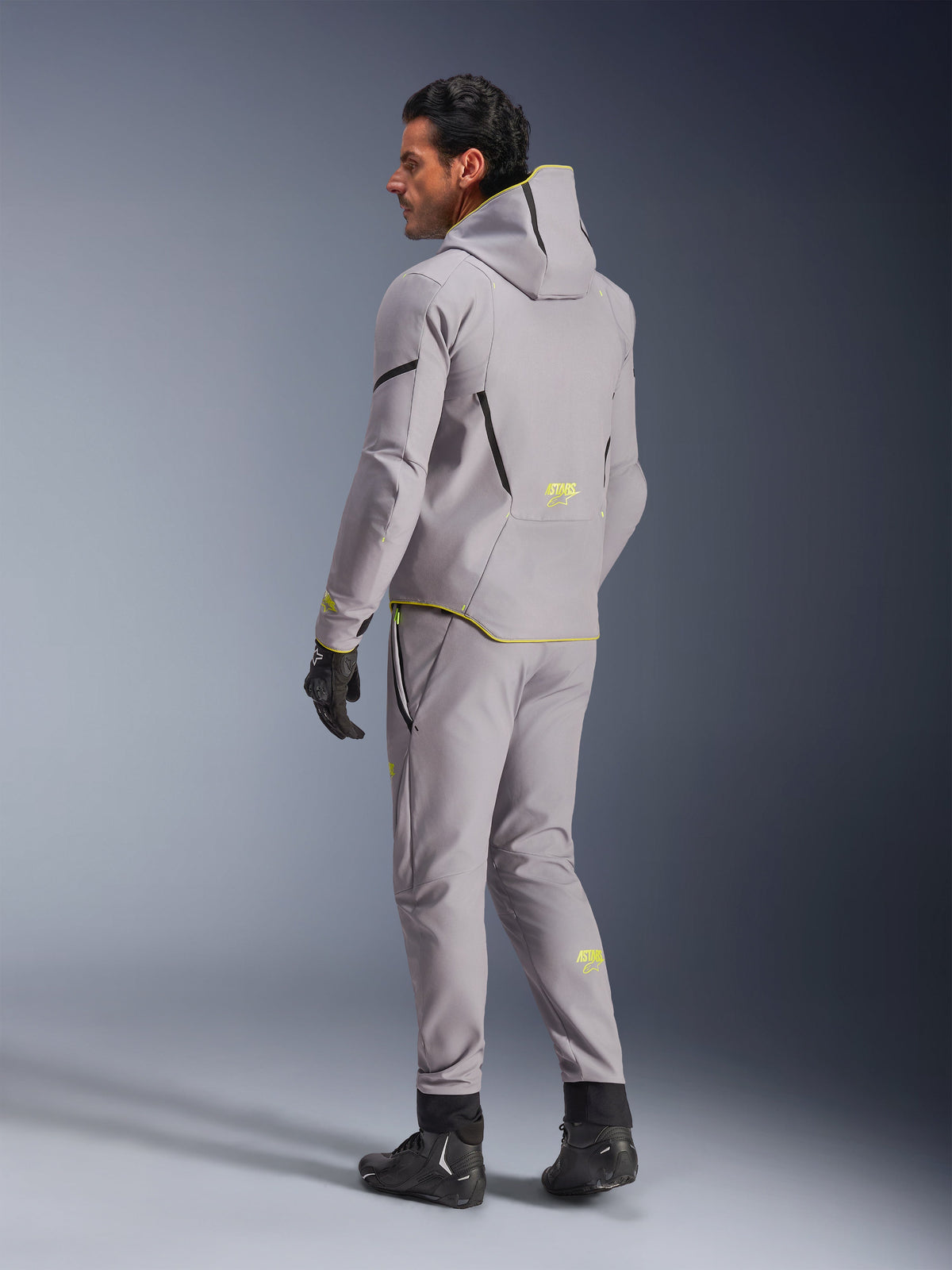 Alpinestars Aeroshell Waterproof Jacket, Mid Gray Yellow Fluo, worn with matching gray pants, featuring a hoodie design with yellow accents and logos, back view, paired with black riding gloves and boots, modern motorcycle apparel