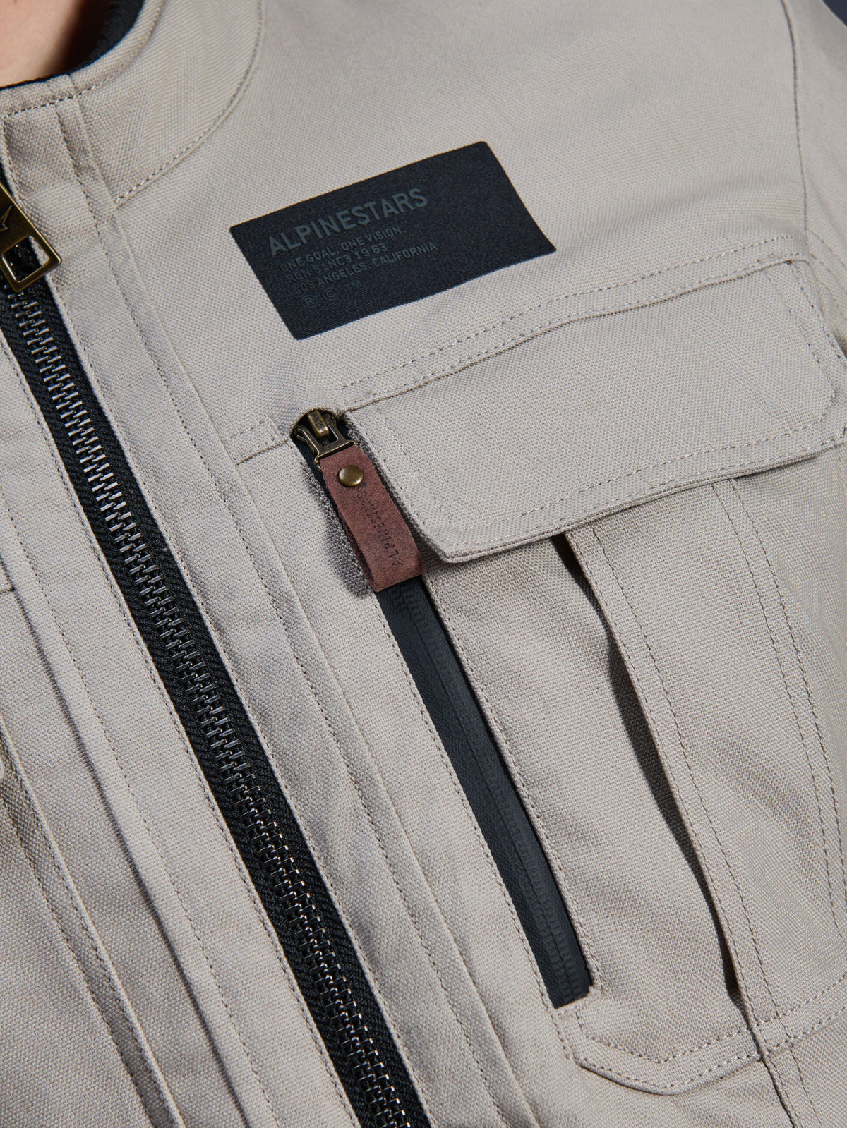 Alpinestars Borrego Drystar® Jas, Warm Gray, detailopname van de borststreek met een zwarte Alpinestars merkpatch, borstzak met klep en een verticale rits met een bruin lederen treklipje, duurzame grijze textiel canvas constructie.