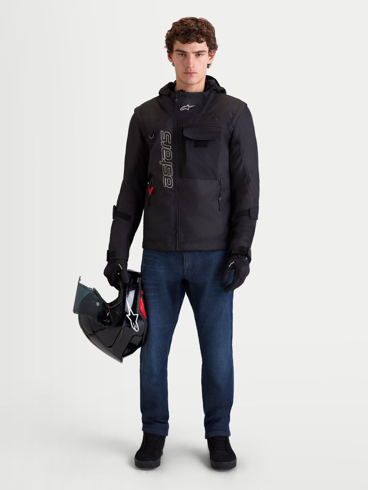 Alpinestars Morfex Parka WP motorjas, zwarte waterdichte urban motortextieljas voor heren, vooraanzicht op model met jeans die een helm vasthoudt, met modulair design en geïntegreerde capuchon