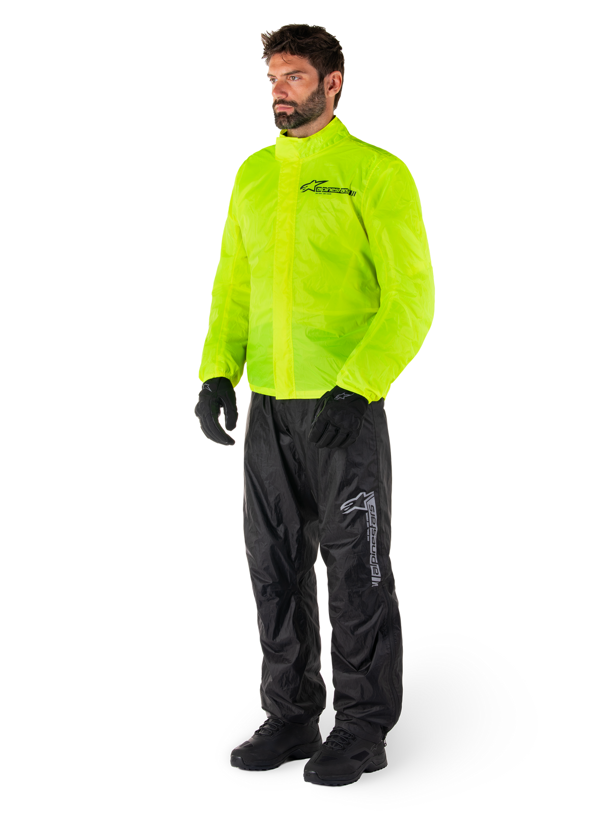 Man draagt Alpinestars Hurricane Rain V2-jas in Yellow Fluo en Hurricane Rain V2-broek in het zwart, goed zichtbare waterdichte motorregenkleding, voorzien van reflecterende logo's op de borst en het bovenbeen, complete outfit met zwarte handschoenen en laarzen.