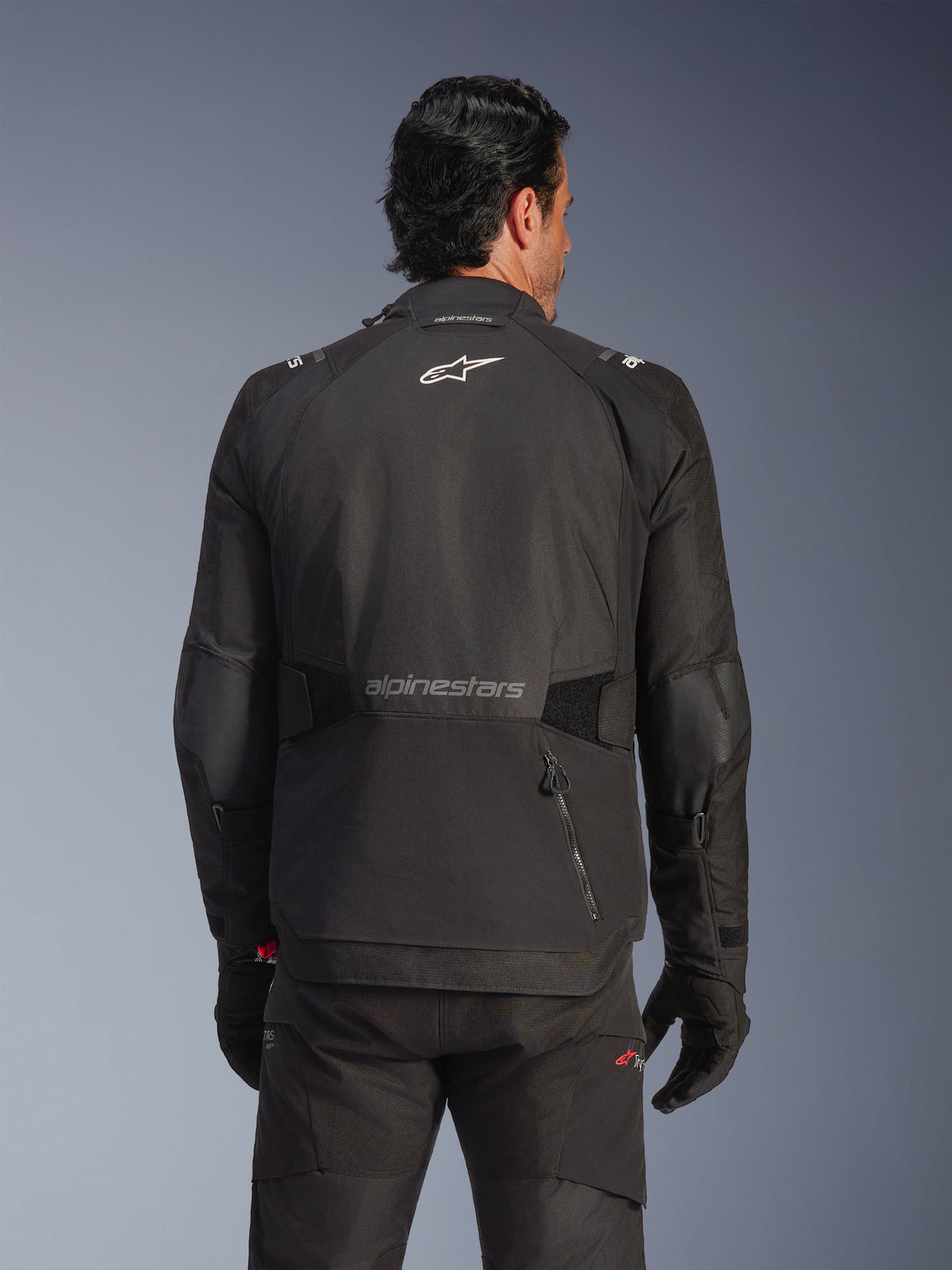 Alpinestars Andes V4 Drystar® Jacket, zwart, achteraanzicht op een model, textiele adventure-motorjas met reflecterende logo's en een achterzak met ritssluiting, gecombineerd met bijpassende zwarte broek en handschoenen
