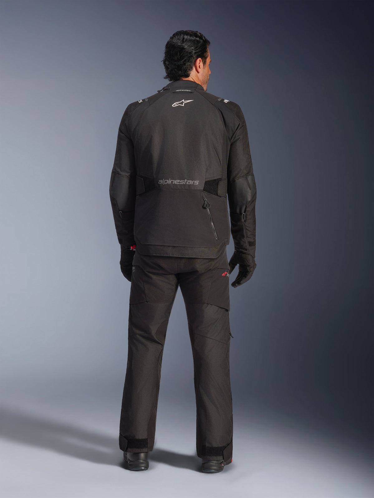 Alpinestars Andes V4 Drystar® Jas in het zwart, achteraanzicht van een mannelijke motorrijder met bijpassende motorbroek en handschoenen, Alpinestars-logo en woordmerk op de rug, technische all-weather adventure-uitrusting
