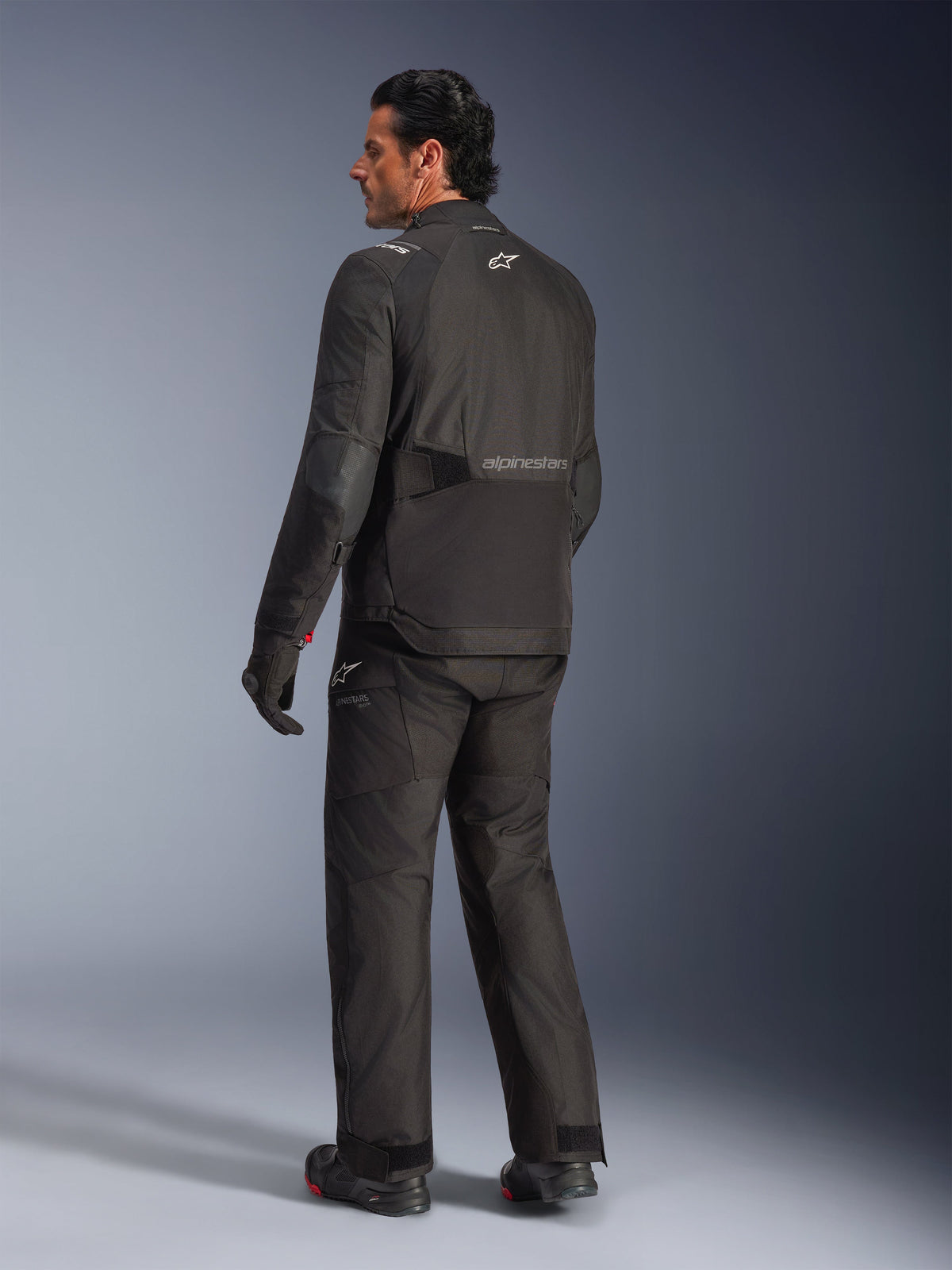 Alpinestars Andes V4 Drystar® jas, zwart, achteraanzicht van rijder in complete motoruitrusting, textiele adventure-jas met Alpinestars-logo op de rug, gedragen met bijpassende zwarte broek en laarzen, all-weather road en adventure rijstijl