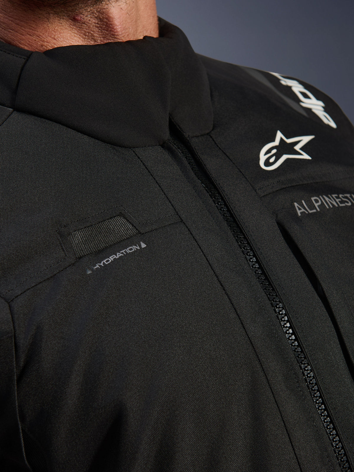 Alpinestars Andes V4 Drystar® Jas, zwart, close-up van borstdetail met lus voor de hydratatieslang, waterdichte VISLON®-voorritssluiting en Alpinestars-logo op duurzaam textielmateriaal