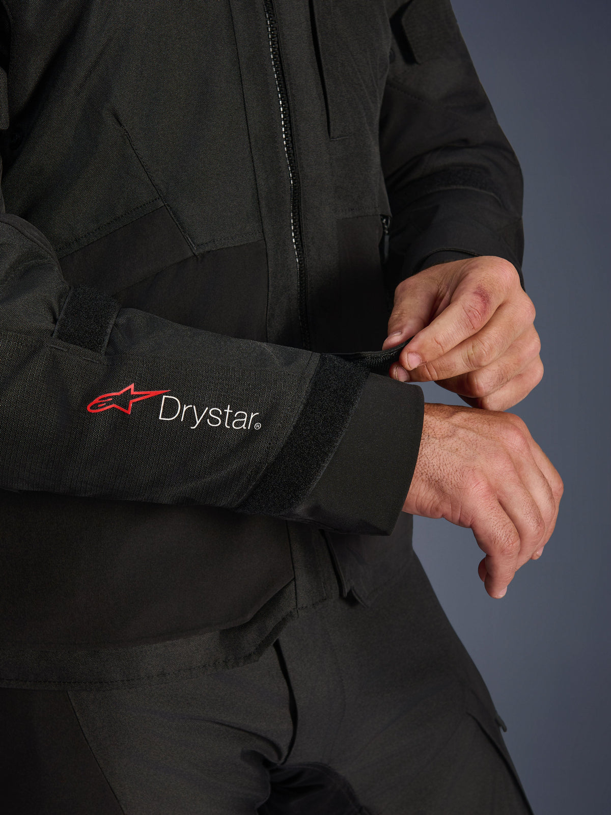 Alpinestars Andes V4 Drystar® Jas, zwarte textiele motorjas, close-up van de mouw met rood Alpinestars-logo en witte Drystar®-branding, motorrijder die de velcro polssluiting verstelt voor een goede pasvorm, functioneel adventure detail