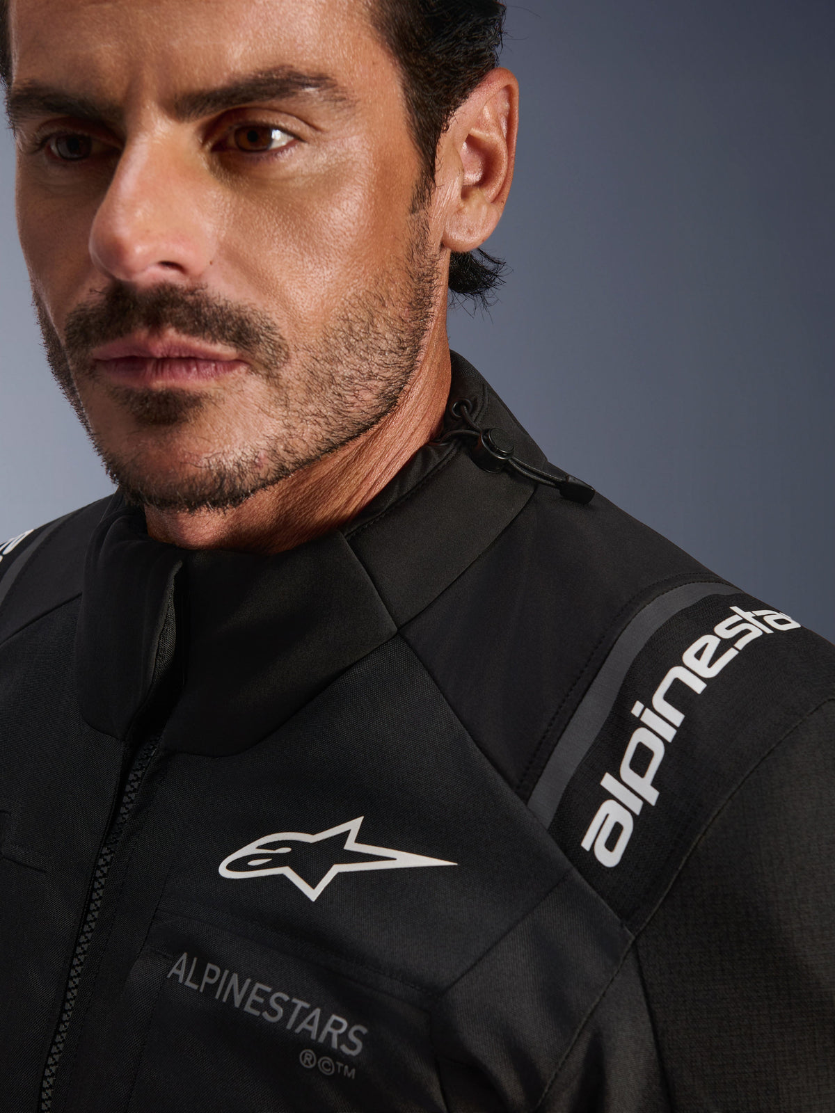 Alpinestars Andes V4 Drystar® Jacket, zwarte textiele motorjas, close-up detail gedragen door een man, voorzien van witte Alpinestars logo's op de borst en schouder, verstelbaar trekkoord bij de kraag en hoogwaardig adventure-touring ontwerp