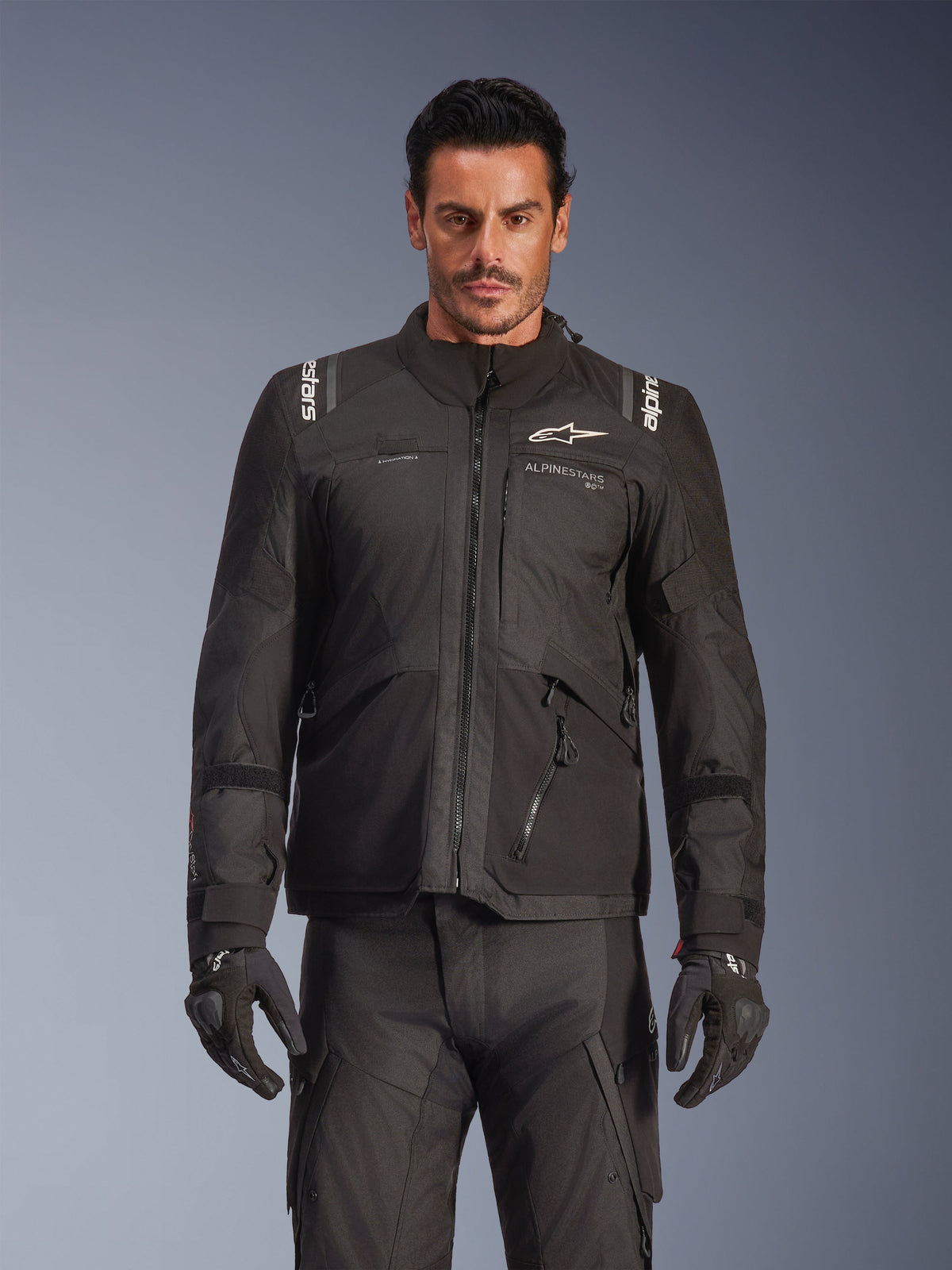 Mannelijk model in de Alpinestars Andes V4 Drystar® Jacket in het zwart, adventure touring motorjas, gecombineerd met bijpassende zwarte motorbroek en handschoenen, voorzien van waterdichte ritsen en witte Alpinestars logo's op de borst en schouders, studio-opname van motorkleding voor alle weersomstandigheden.