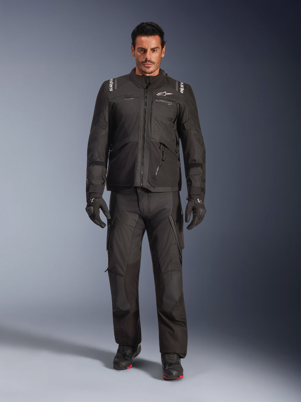 Mannelijk model draagt de Alpinestars Andes V4 Drystar® Jas in het zwart, gecombineerd met een bijpassende adventure motorbroek, zwarte handschoenen en zwarte laarzen, full-body studio-opname van technische touringuitrusting voor alle weersomstandigheden met Alpinestars-branding.