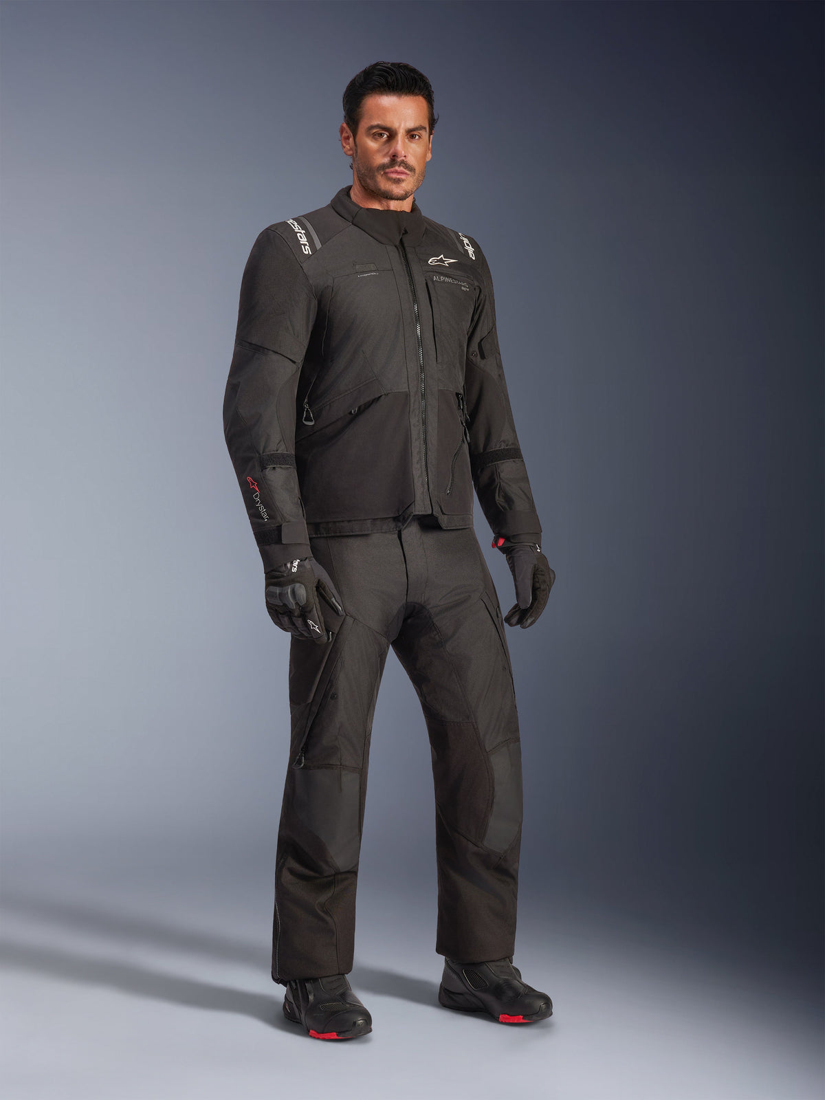 Alpinestars Andes V4 Drystar® Jas, zwarte motorkledingset gedragen door een model, adventure-touring textiele jas en broek, zwarte handschoenen en laarzen, studio-opname, strak modern ontwerp voor rijden in alle weersomstandigheden