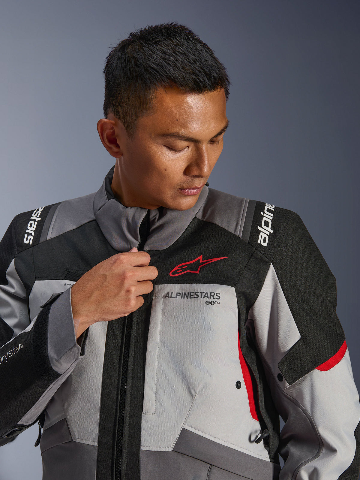 Model draagt Alpinestars Andes V4 Drystar® Jacket, donkergrijs en ijsgrijs met zwarte en felrode accenten, verstelt de rits aan de voorzijde, met het rode Astars-logo en Drystar®-branding, adventure touring-stijl