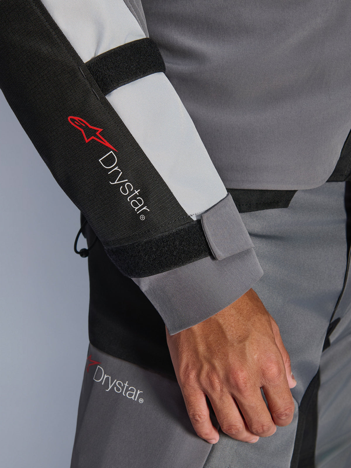 Close-up van de mouw van de Alpinestars Andes V4 Drystar® jas, donkergrijs en ijsgrijs met zwarte accenten, voorzien van het Drystar®-logo en verstelbare onderarmriemen, onderdeel van een waterdichte textiele adventure motoroutfit