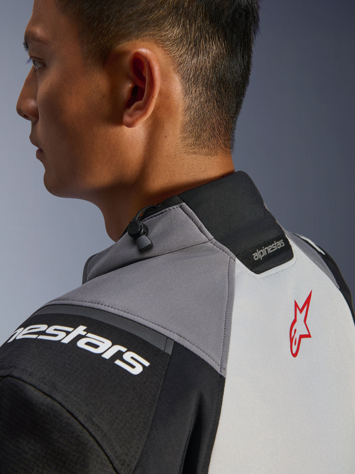 Alpinestars Andes V4 Drystar® Jas, close-up van de kraag en de bovenrug, donkergrijs en ijsgrijs met zwarte accenten, rood sterlogo, voorzien van een trekkoordversteller bij de kraag en Alpinestars-branding, duurzame textielconstructie
