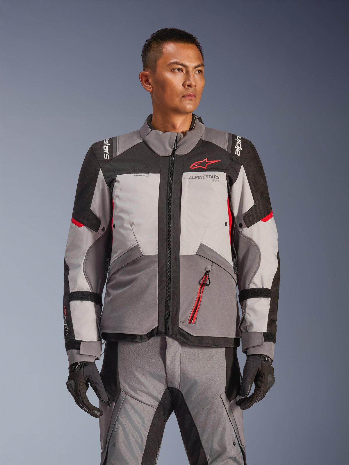 Alpinestars Andes V4 Drystar® jas, adventure touring motorjas, donkergrijs en lichtgrijs met zwarte en felrode accenten, gedragen met bijpassende broek en handschoenen, waterdicht en ademend ontwerp voor adventure ritten onder alle weersomstandigheden