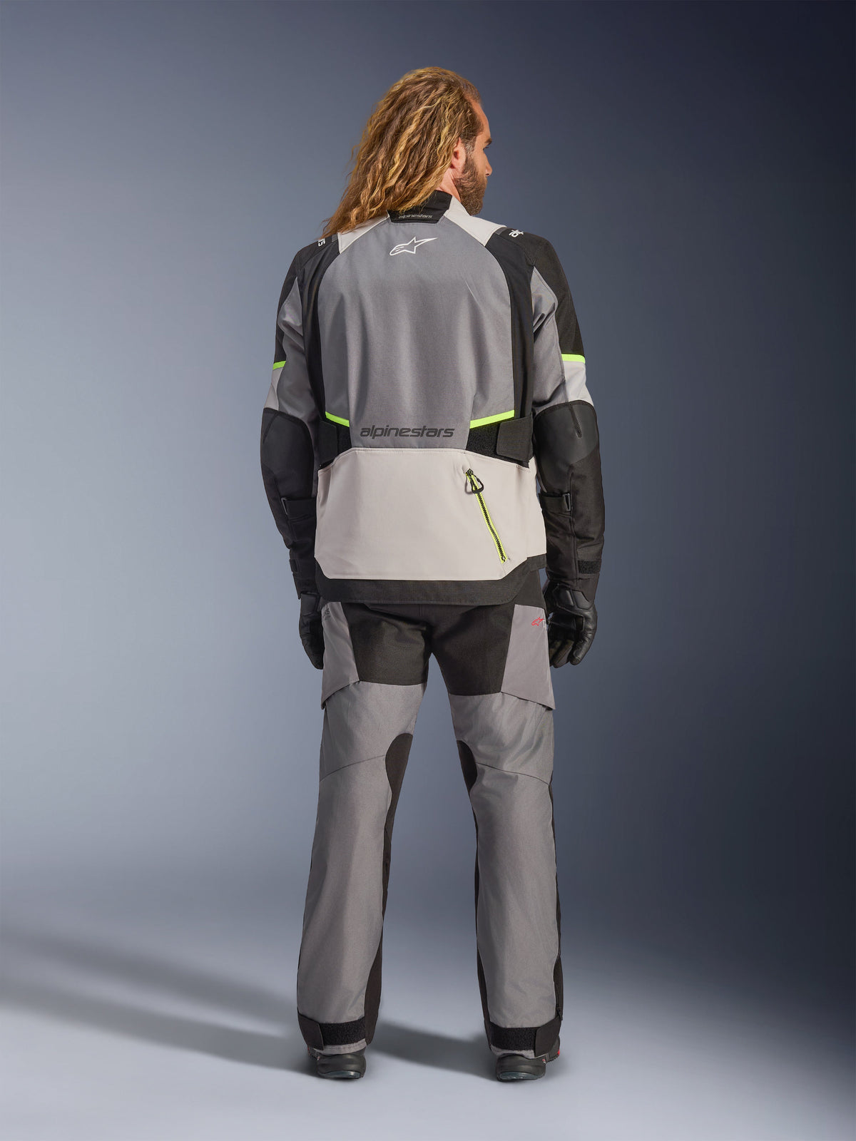 Alpinestars Andes V4 Drystar® Jas, adventure motorjas, donkergrijs en ijsgrijs met zwarte en fluo gele accenten, achteraanzicht gedragen met bijpassende broek en zwarte handschoenen, voorzien van een grote achterzak met gele rits en verstevigde schouder- en elleboogpanelen