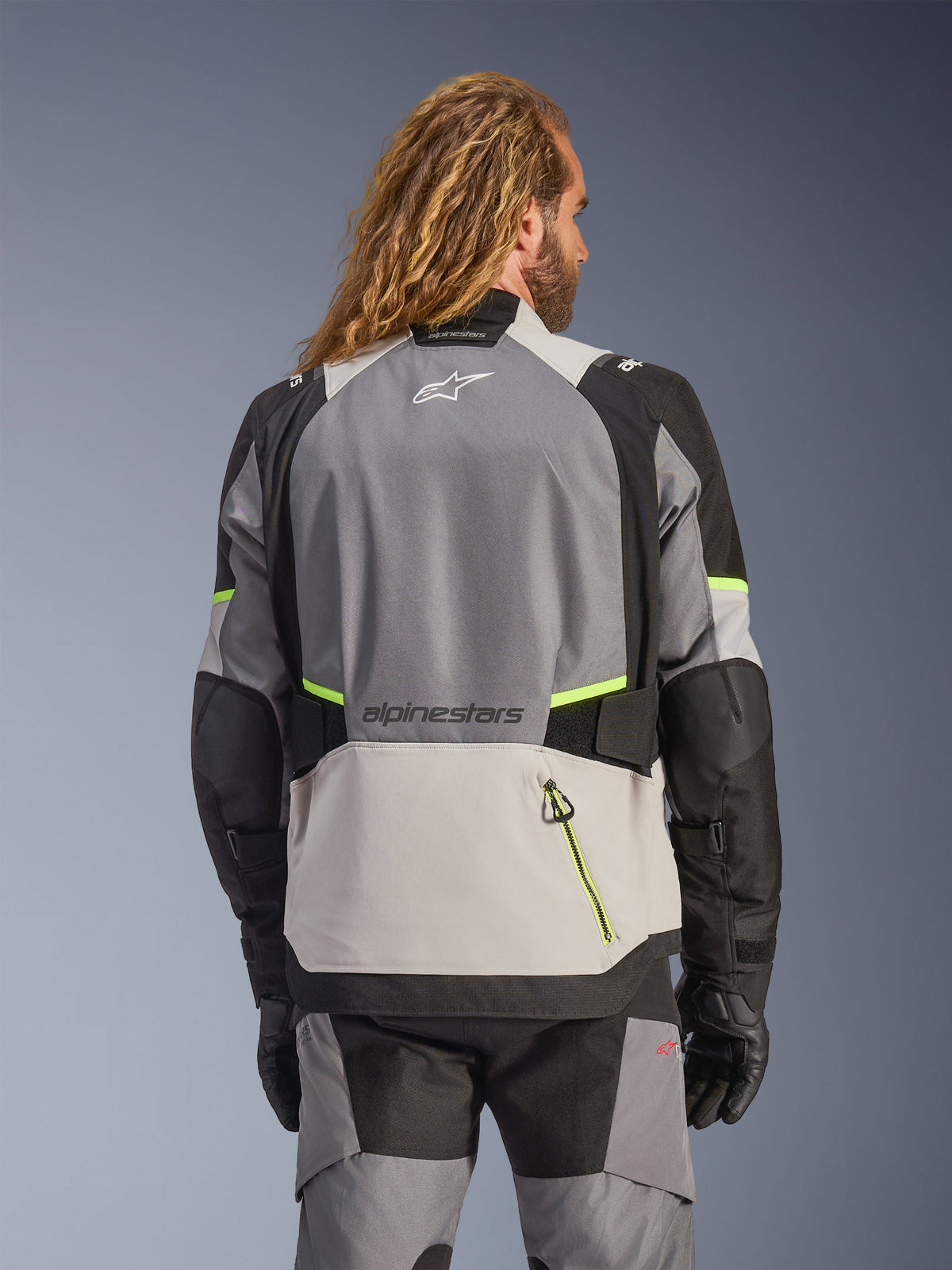 Alpinestars Andes V4 Drystar® Jas, achteraanzicht van adventure motorjas in donkergrijs, ijsgrijs en zwart met fluorescerend gele fluo-accenten, voorzien van versterkte textielpanelen en Alpinestars-branding, gedragen met bijpassende motorbroek en zwarte handschoenen