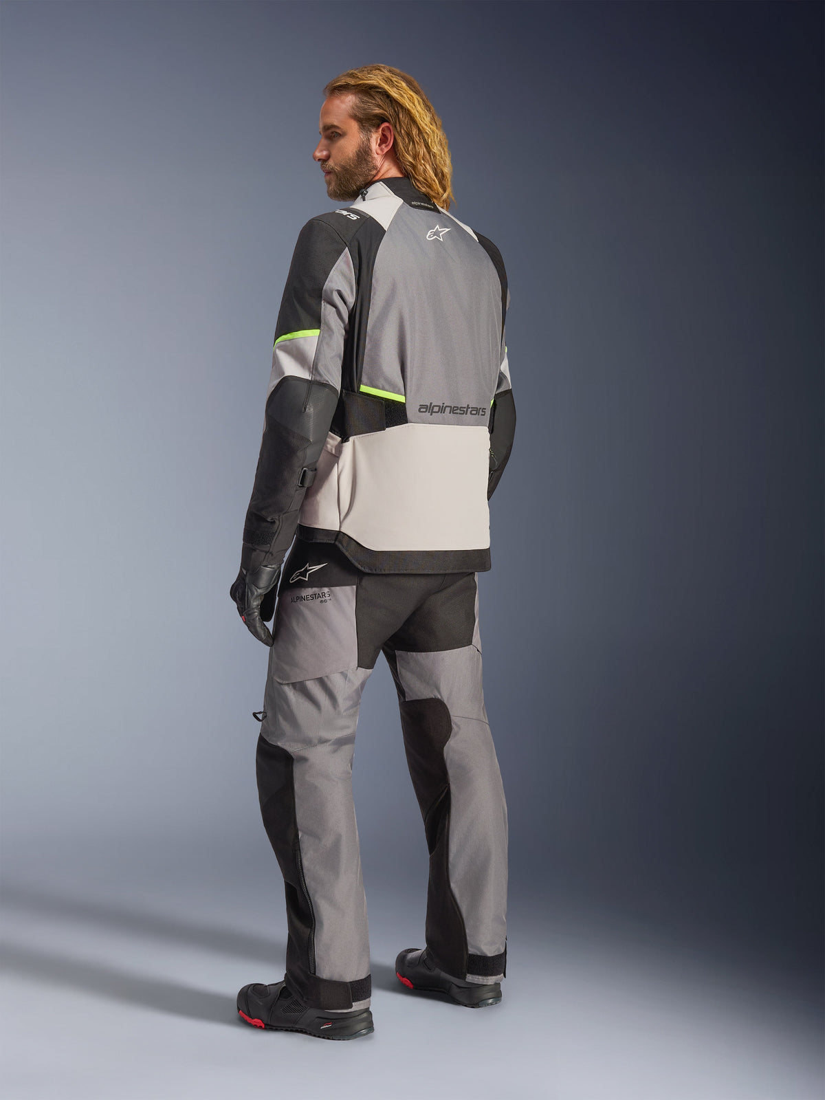 Alpinestars Andes V4 Drystar® Jas, donkergrijs, lichtgrijs, zwart en fluor geel, achteraanzicht van motorrijder die een bijpassende adventure touring broek en laarzen draagt, voorzien van versterkte grijze textielpanelen, neongele accenten en Alpinestars-branding, motoruitrusting voor alle weersomstandigheden.