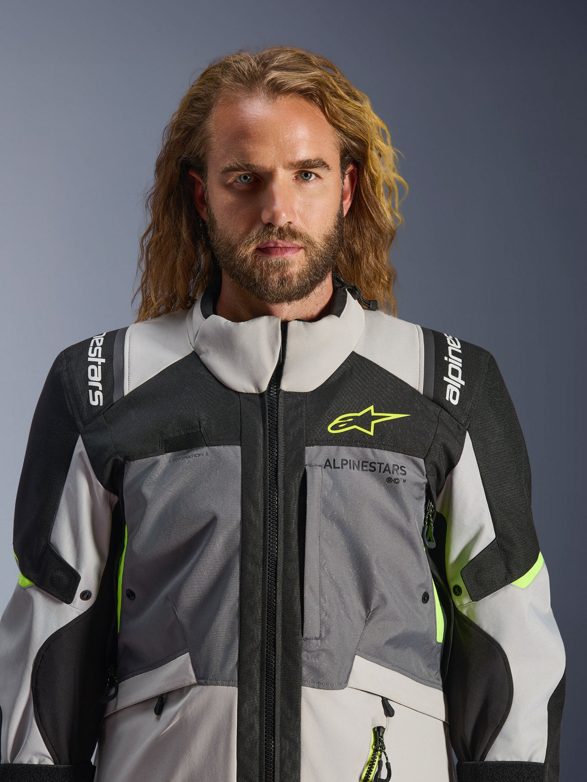 Alpinestars Andes V4 Drystar® Jas, donkergrijs, ijsgrijs, zwart en fluogeel, gedragen door een man met lang golvend haar, voorzien van technische multi-fabric constructie, Alpinestars-branding op de borst en schouders, hoog kraagontwerp voor adventure-rijden