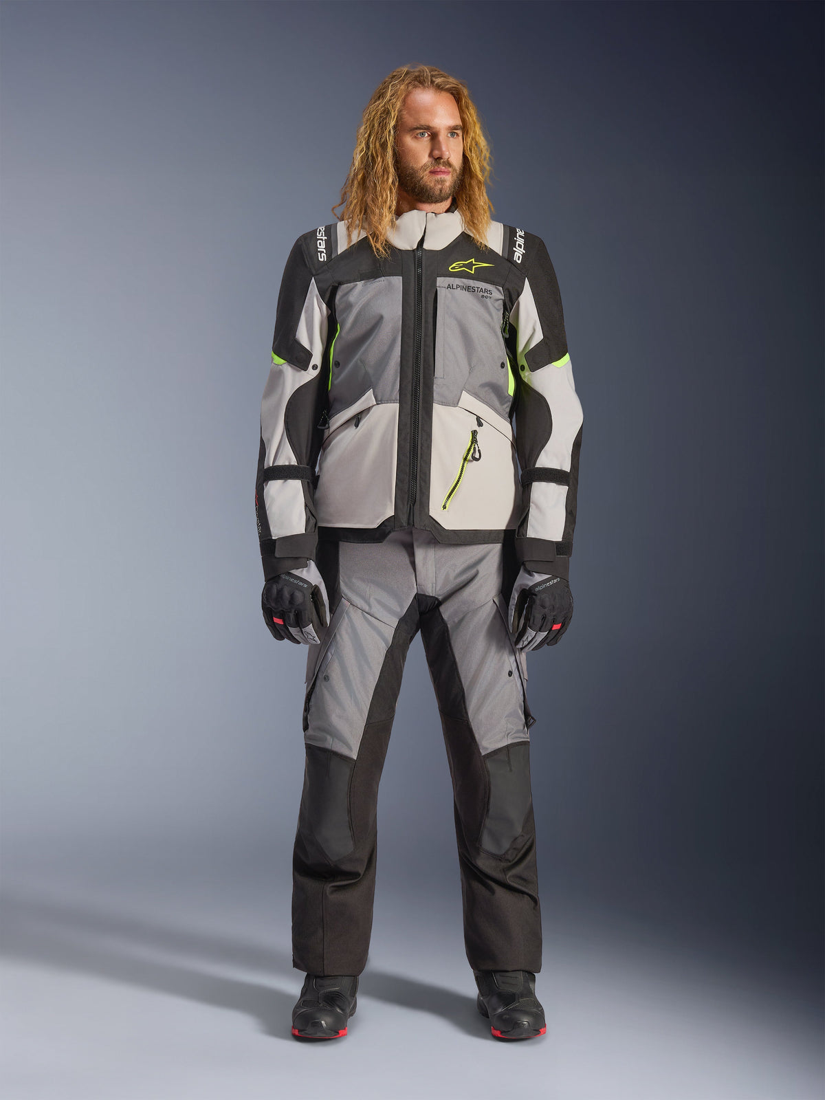 Mannelijk model met lang blond haar in de Andes V4 Drystar® Jas in Dark Gray, Ice Gray, Black en Yellow Fluo, gecombineerd met een bijpassende adventure motorbroek en zwarte laarzen, een volledige technische motoruitrusting voor touring in alle weersomstandigheden.