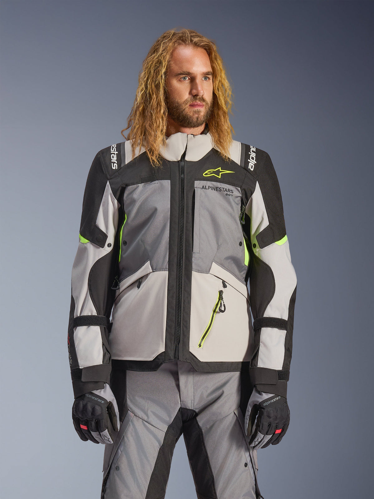 Alpinestars Andes V4 Drystar motorjas, adventure touring motorjas, donkergrijs en lichtgrijs met neongele accenten, gedragen door een model met bijpassende broek en handschoenen, textielontwerp met meerdere zakken voor rijden in alle weersomstandigheden