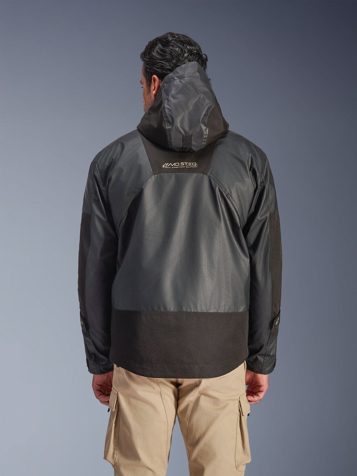 Achteraanzicht van een man die de zwarte Alpinestars Mospher Waterproof Jacket draagt, voorzien van een capuchon en duurzame ripstop textielconstructie, gecombineerd met een beige cargobroek, met nadruk op het MO.ST.EQ.-logo op de bovenrug, urban motorjas ontwerp.