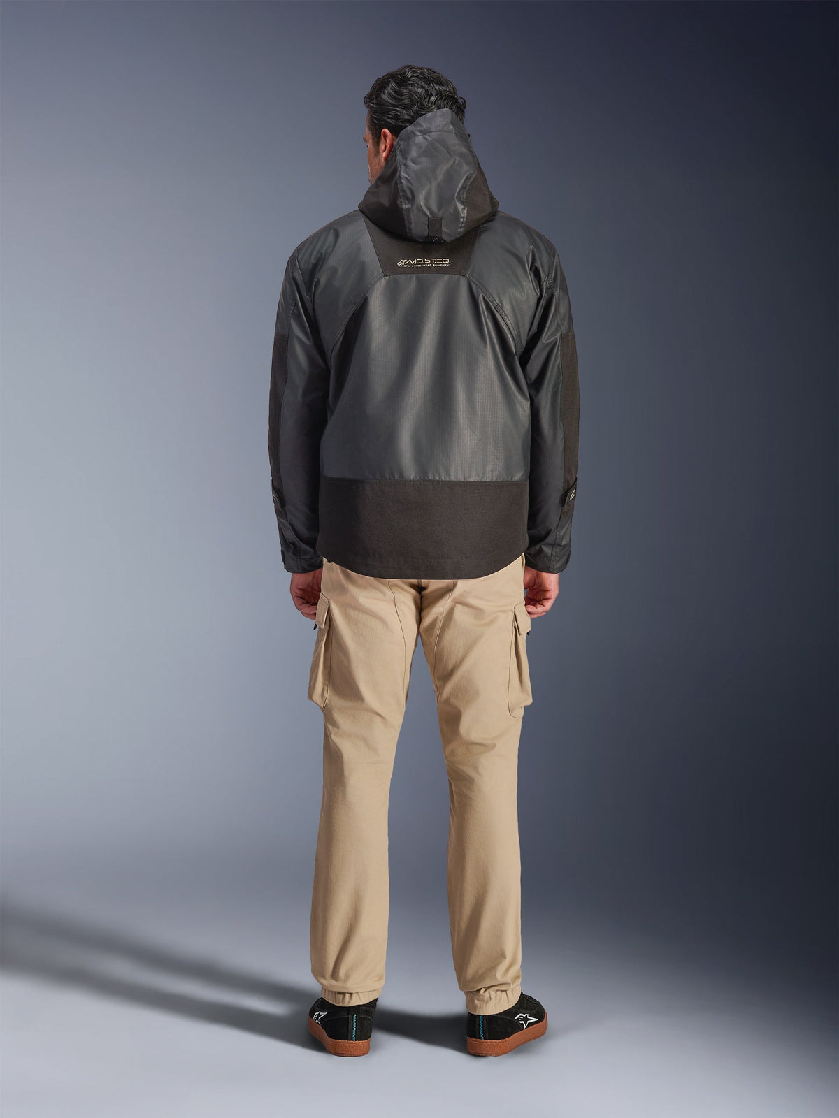 Alpinestars Mospher Waterproof Jacket, zwarte motorparka van achteren gefotografeerd, met MO.ST.EQ-logo op de bovenrug en ripstop-stofconstructie, gedragen met een beige cargobroek en zwarte sneakers, urban commuter-stijl.