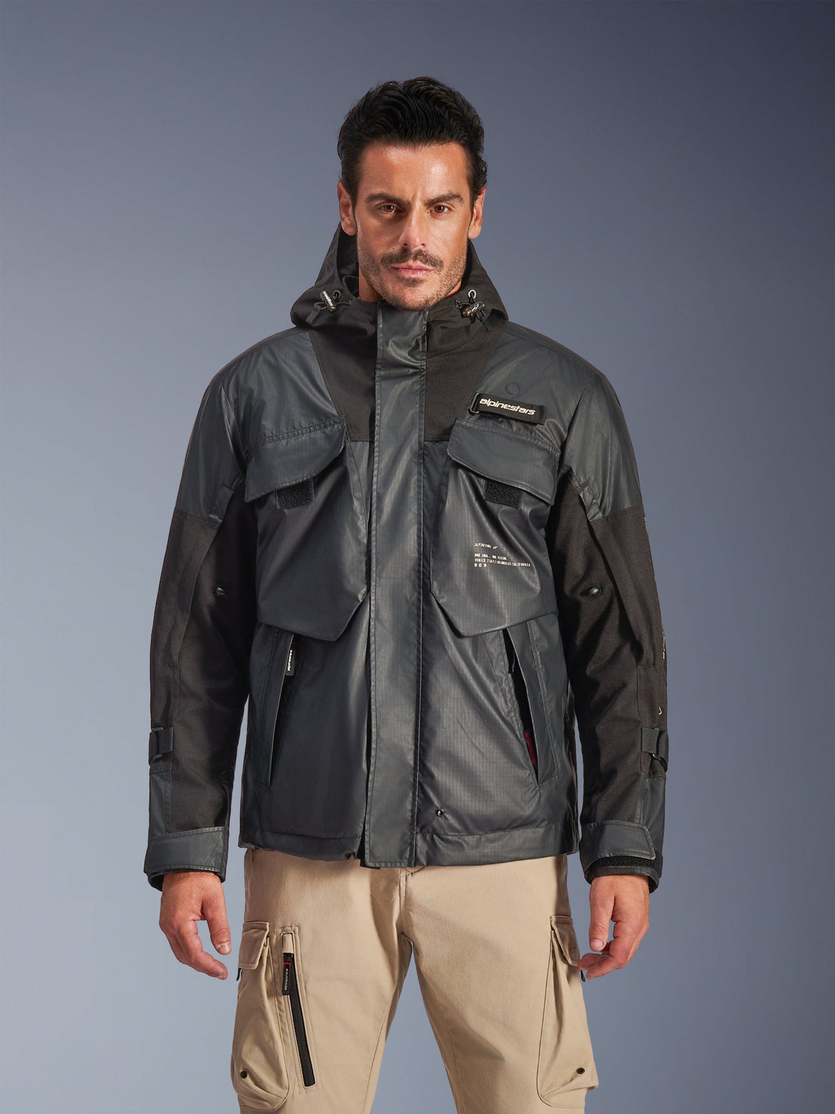 Alpinestars Mospher waterdichte jas, urban motorparka voor heren, zwart en donkergrijs ripstop-materiaal, ontwerp met capuchon en meerdere grote functionele zakken, gedragen door een model met een kaki cargobroek voor een tactische look