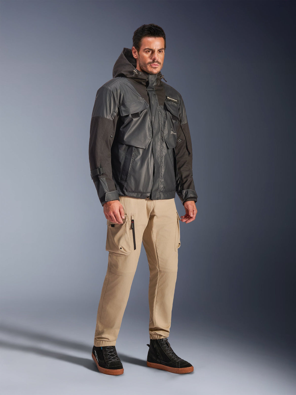 Mannelijk model draagt de Alpinestars Mospher Waterproof Jacket in het zwart, een op utility geïnspireerde waterdichte parka met een multi-pocket design en capuchon, gecombineerd met een beige cargobroek en zwarte high-top sneakers, stedelijke motoruitrusting voor woon-werkverkeer.