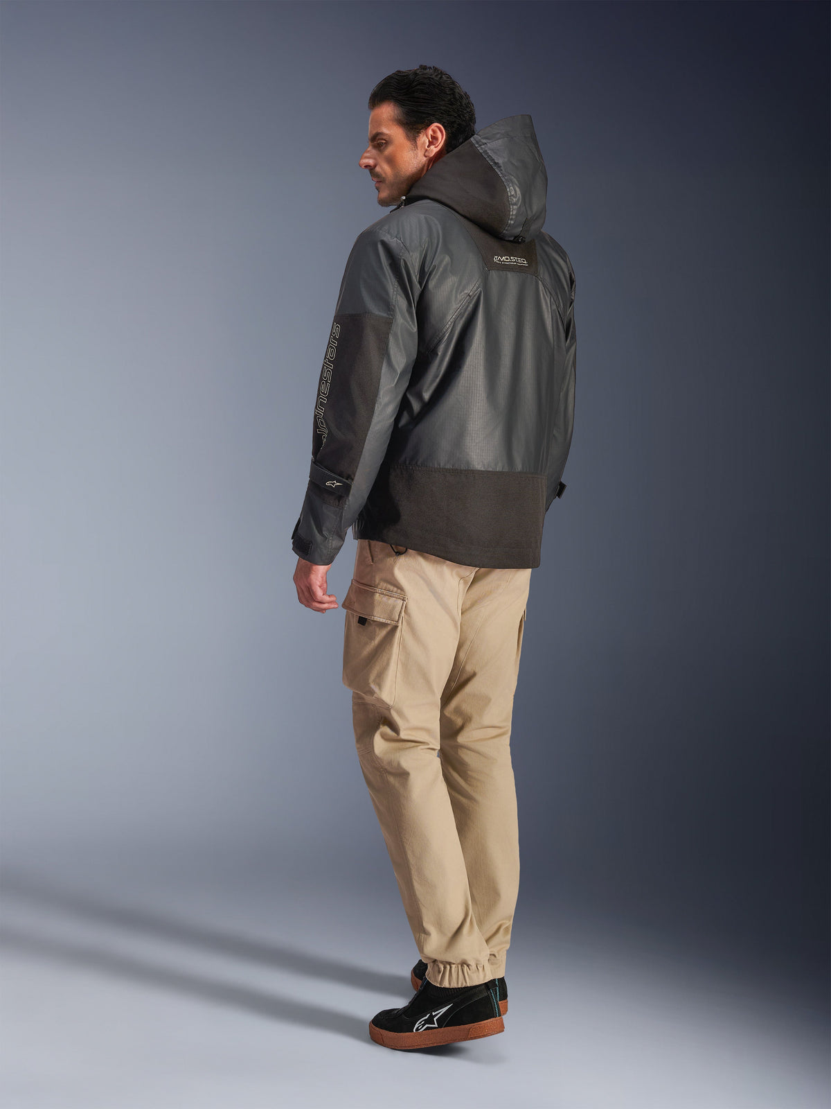 Alpinestars Mospher Waterproof Jacket in het zwart, man in achteraanzicht en zijprofiel, gecombineerd met een beige cargobroek en zwarte sneakers, parka-ontwerp met capuchon, ripstop-stof, Alpinestars-logo op de mouw, moderne urban motorstijl.
