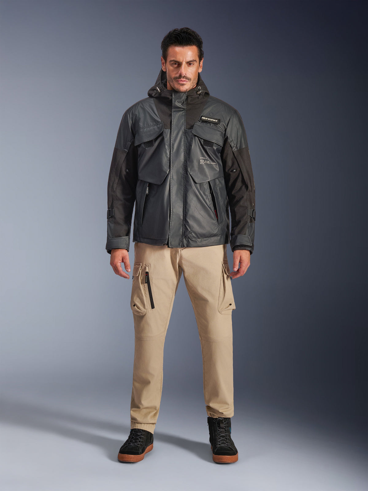 Mannelijk model in de Alpinestars Mospher Waterproof Jacket in Black, urban eigentijdse motorkleding, tactisch ontwerp met meerdere functionele zakken en capuchon, gecombineerd met een beige cargobroek en zwarte schoenen, full-body studioportret