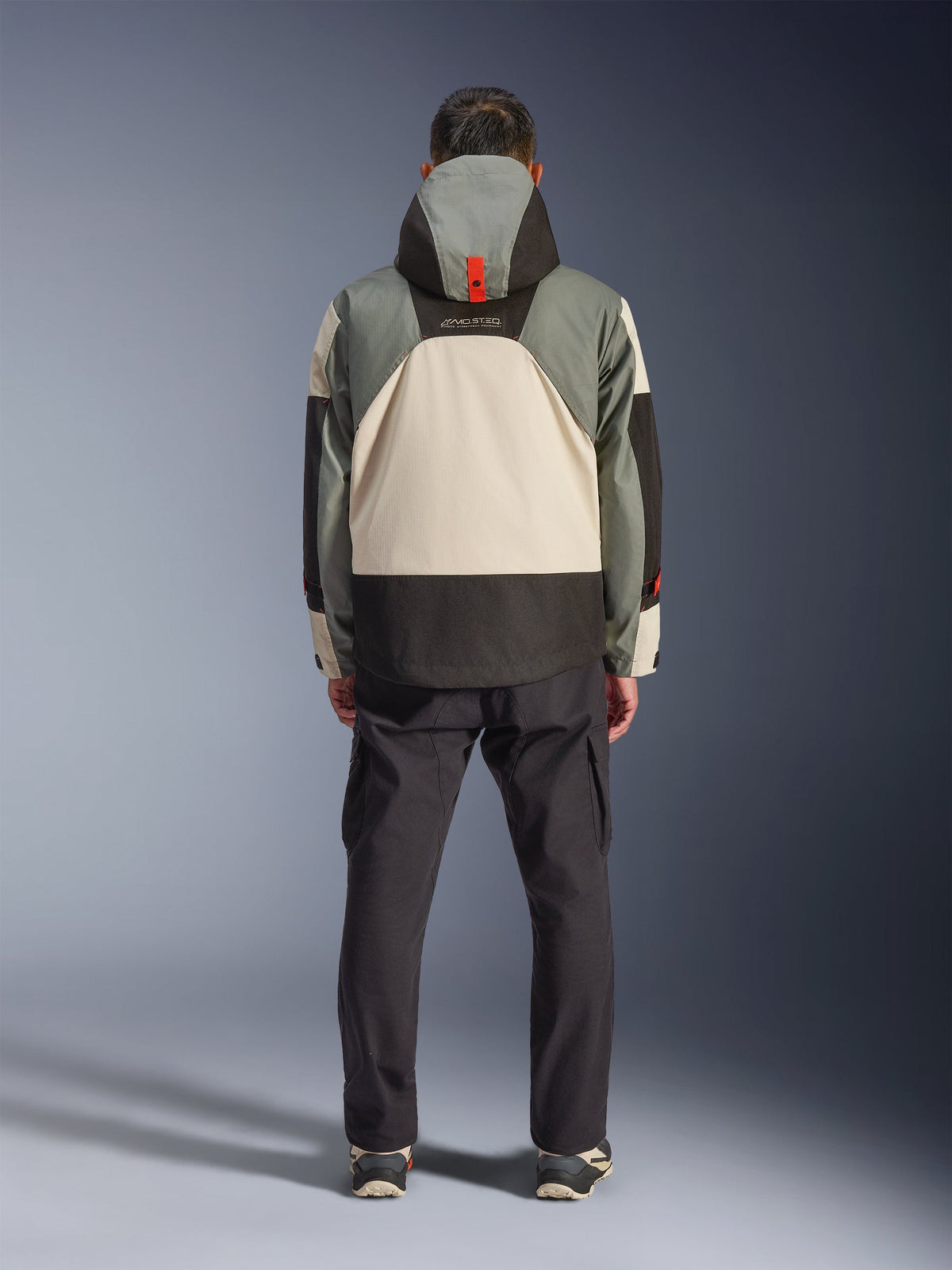 Achteraanzicht van een rijder die de Alpinestars Mospher Waterproof Jacket in Fur Gray Black Ice draagt, tactisch geïnspireerde parka met grijs, zwart en crème color-blocking, voorzien van een capuchon met oranje banddetail, gecombineerd met een zwarte cargobroek.