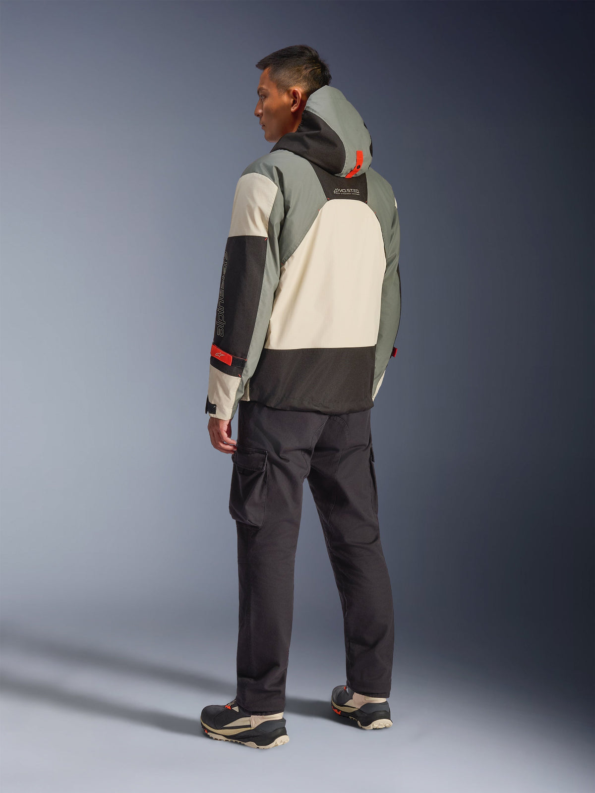 Alpinestars Mospher Waterproof Jas in Fur Gray Black Ice, achteraanzicht van een model dat een tactische motorparka draagt met crème, grijze en zwarte panelen, voorzien van rode accenten en een capuchon, gecombineerd met een zwarte cargobroek en technische laarzen