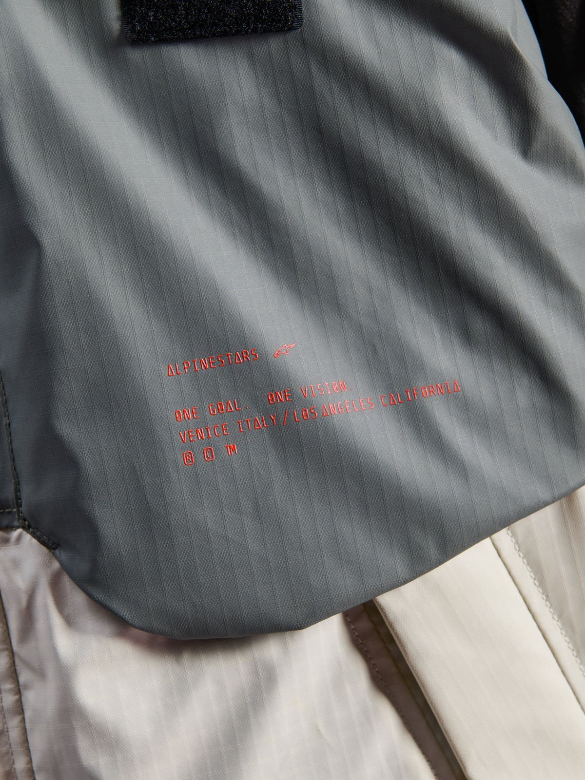 Alpinestars Mospher Waterproof Jacket, motorparka, Fur Gray en Black Ice, detail van technische ripstop-stof met rode Alpinestars-branding en slogan, nadruk op duurzame constructie en urban tactisch design