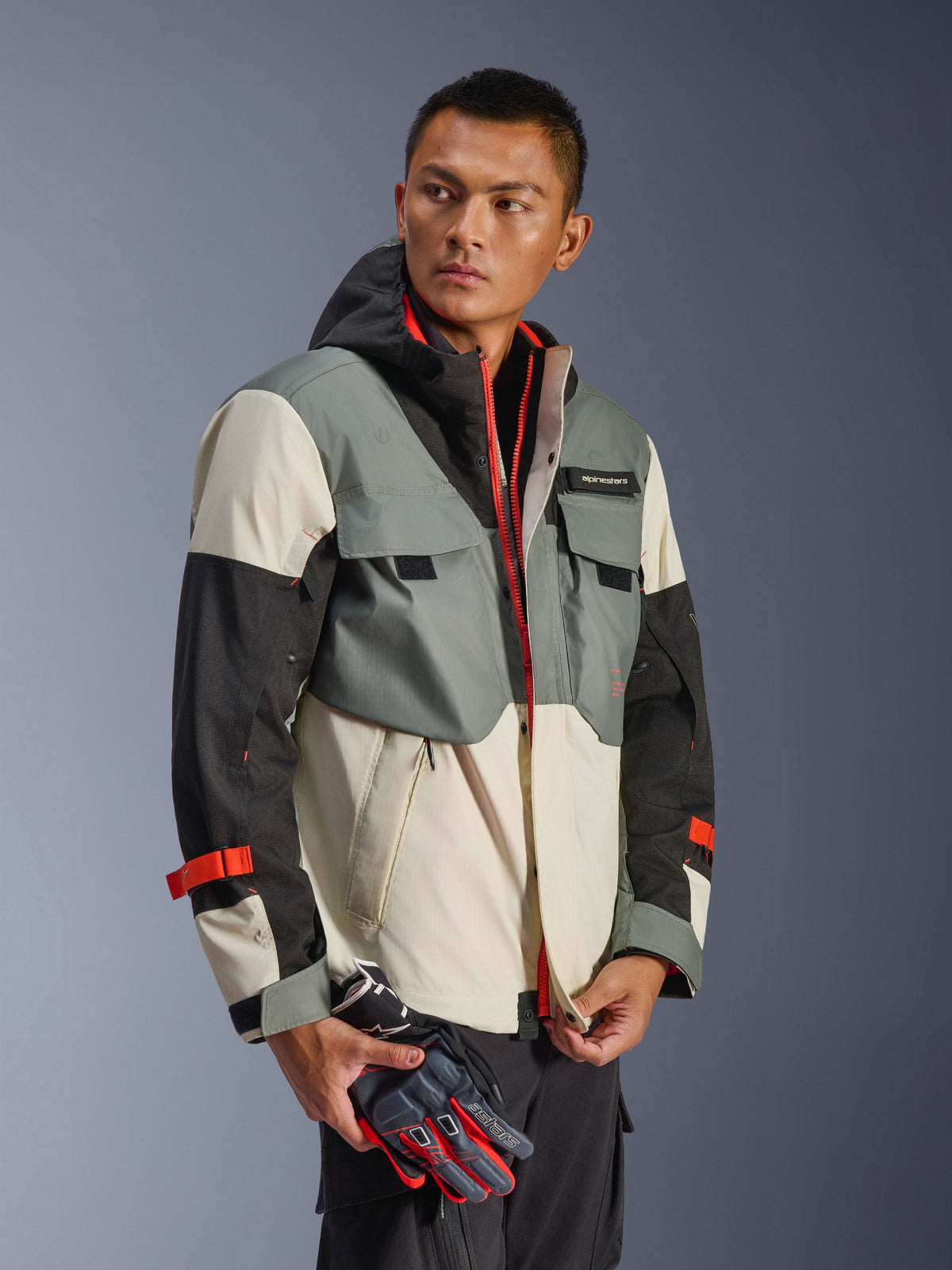 Alpinestars Mospher Waterproof Jacket in Fur Gray Black Ice, multi-paneel textiele parka met grijze, zwarte en off-white vlakken, voorzien van felrode ritsen en bandaccenten, tactisch geïnspireerd ontwerp met capuchon en functionele zakken, gedragen door een model dat motorhandschoenen vasthoudt.