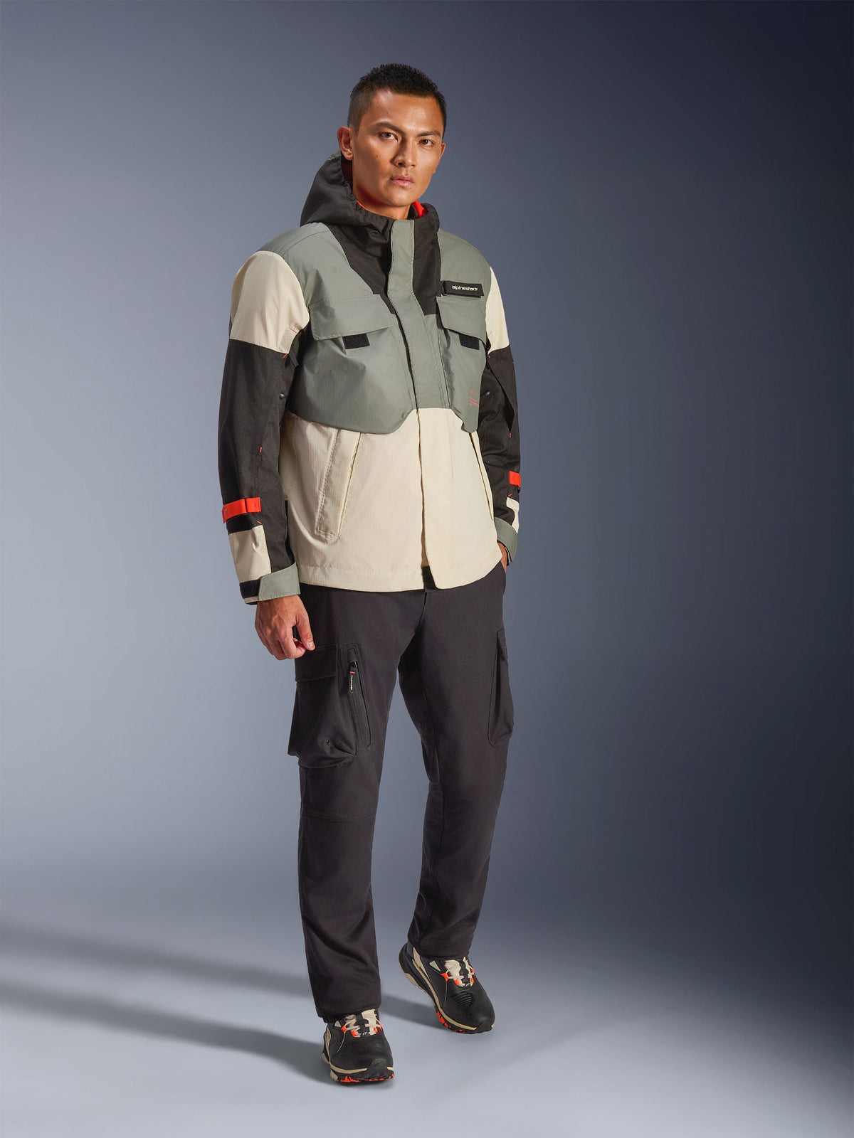Alpinestars Mospher Waterproof Jas in Fur Gray Black Ice, meerkleurige technische motorparka met grijze, zwarte en gebroken witte panelen en rode accenten, functioneel ontwerp met grote zakken en capuchon, gedragen met donkergrijze cargobroek en zwarte sneakers, urban contemporary motoruitrusting