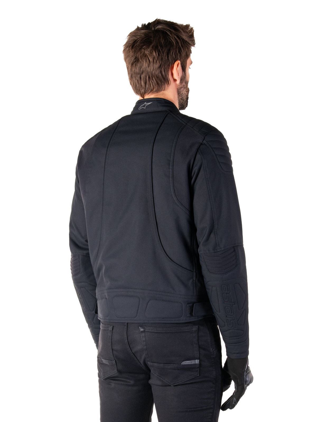 Alpinestars Clayton WR jas in het zwart, driekwart achteraanzicht op een mannelijk model, urban motorjas met gewatteerde vulling op de schouders en ellebogen, technische stiklijnen en reflecterend logo op de kraag, gecombineerd met een zwarte broek en handschoenen.