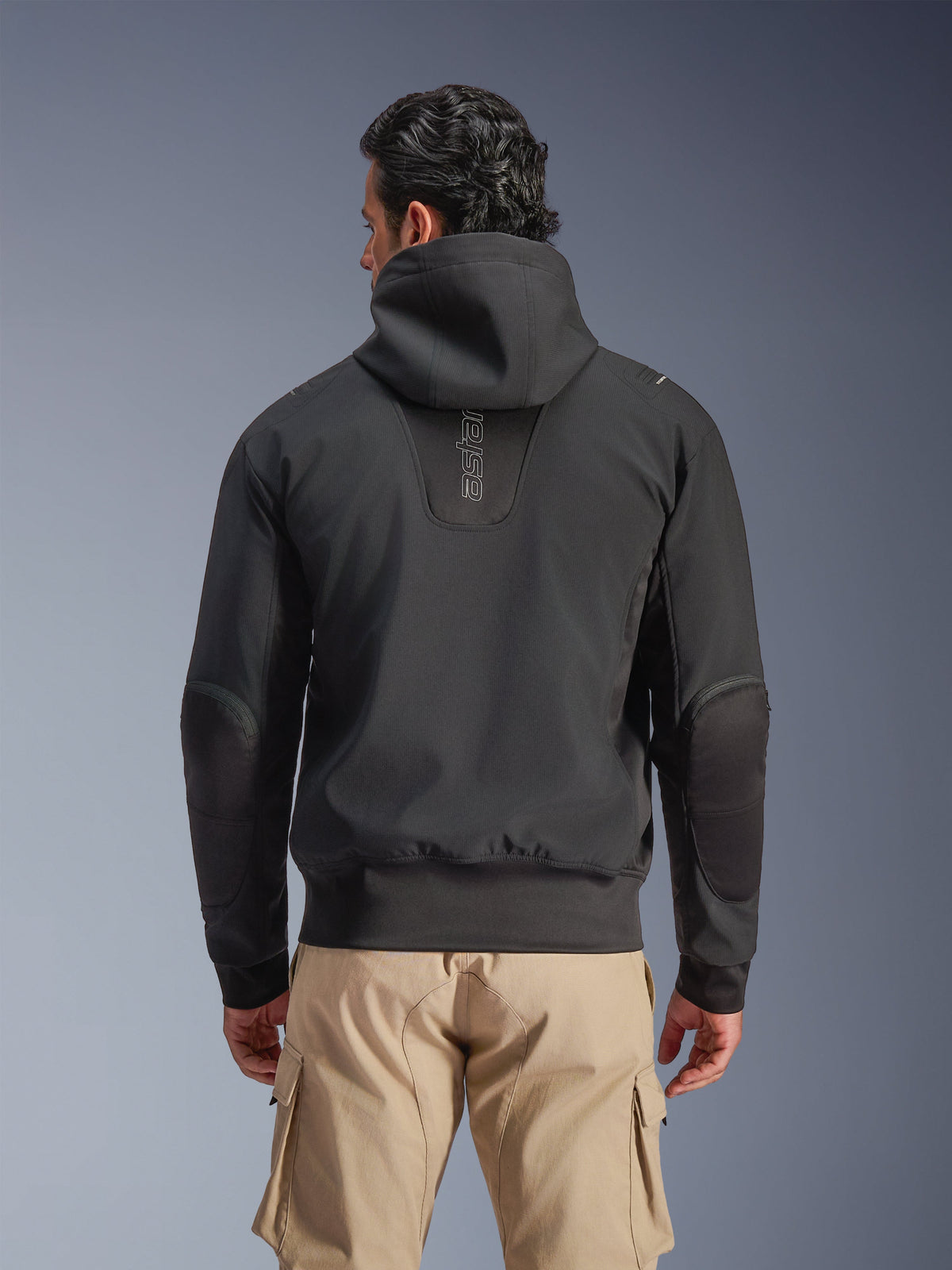 Alpinestars Moblast Waterdichte Jas, zwart, achteraanzicht van stedelijke motorjas met capuchon, voorzien van verticaal 'astars' logo op de rug, elastische taille en manchetten, versterkte ellebogen en modern tech-forward ontwerp, gedragen met beige cargobroek