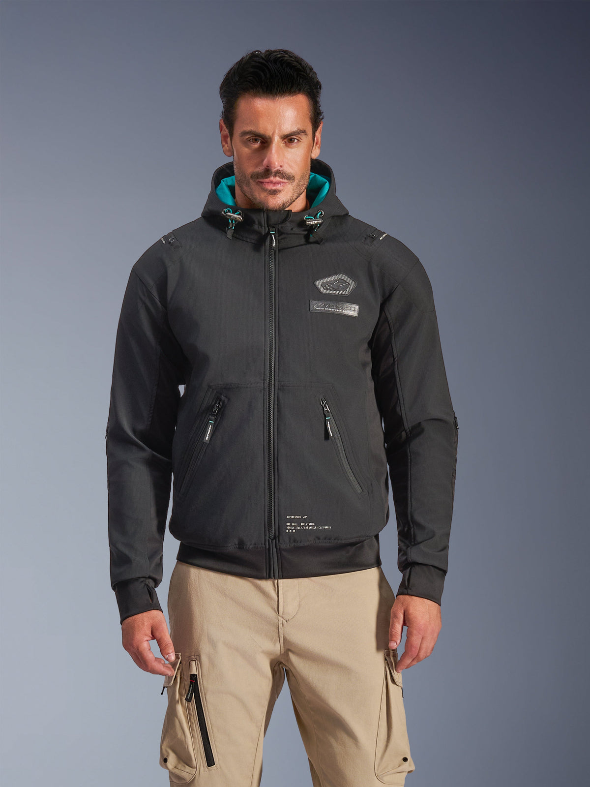 Alpinestars Moblast Waterproof Jacket, urban motorjas voor heren, zwart met turquoise voering in de capuchon, voorzien van 3D-zakken met rits en een ontwerp met capuchon, gedragen met een beige cargobroek, moderne urban motorkleding