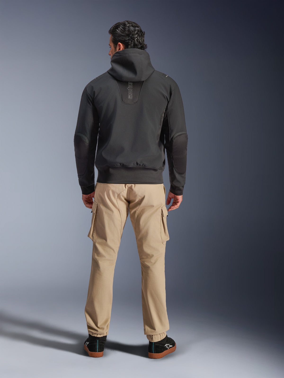 Alpinestars Moblast waterdichte jas, zwart, achteraanzicht van stedelijke motorhoodie, gedragen met beige cargo broek en zwarte sneakers, minimalistisch ontwerp met verticale 'astars' branding op de bovenrug, eigentijdse motorkleding voor alle weersomstandigheden